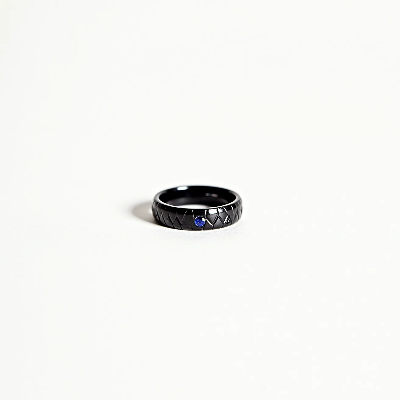 This men's ring features a sleek, dark metal band with a textured pattern resembling a quilted design. At its center, a round-cut blue gemstone is elegantly set, providing a striking contrast to the band’s dark hue. The stone is securely housed in a prong setting, adding both security and sophistication to its appearance. The ring offers a blend of modern aesthetics and timeless elegance, making it a versatile accessory for various occasions.