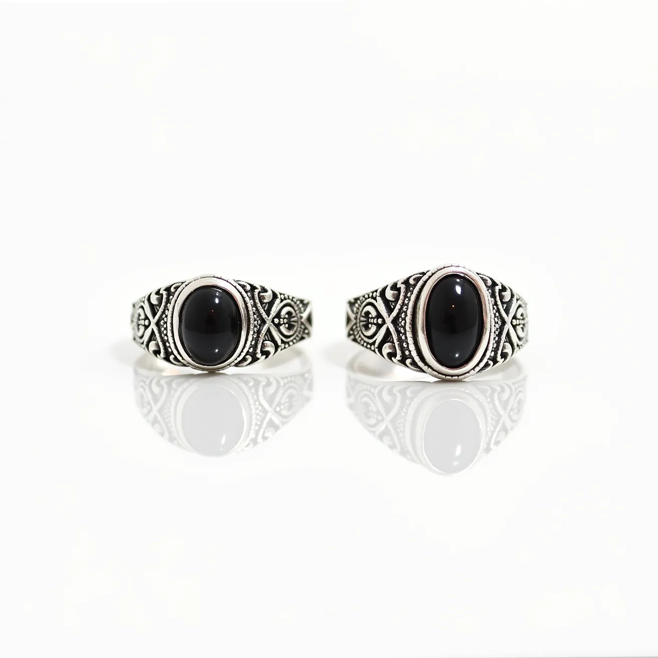 These men's silver rings feature an intricately crafted design with a bold black oval gemstone, likely onyx, as the centerpiece. The stone is set in a bezel setting that securely holds it in place while allowing its smooth, reflective surface to stand out. Surrounding the gemstone, the silver band showcases a detailed pattern with ornamental engravings and textured accents, giving it a striking and classic appearance. The artistry in the design enhances the overall elegance and appeal of the rings.