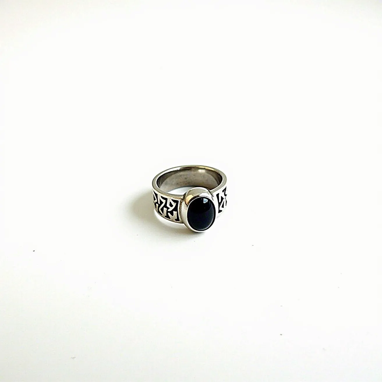 This men's silver ring features an oval-shaped black onyx stone set in a bezel setting. The silver band showcases intricate geometric engravings that add a touch of detail to its bold design. The stone’s smooth, polished surface complements the shine of the silver, creating a striking contrast. There is no clasp or additional attachments, allowing the ring to present a smooth finish overall.