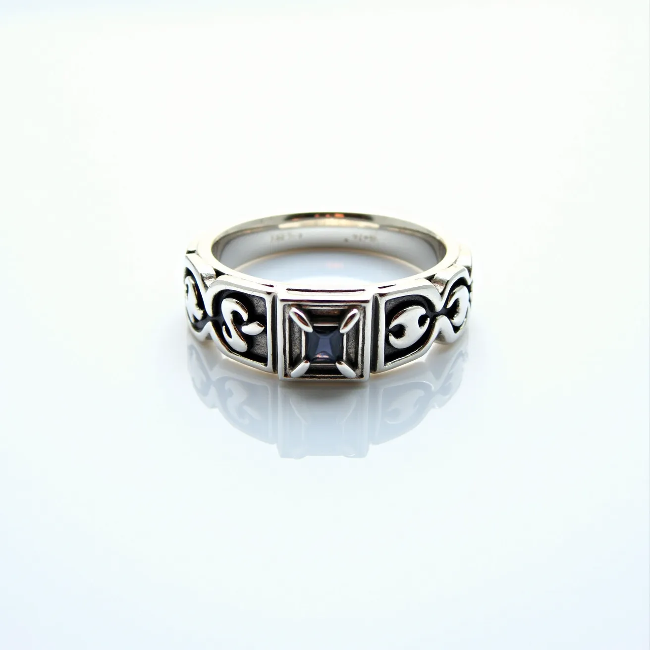 This men's silver ring features an intricately designed band with a striking black square gemstone set in the center. The gemstone is cut in a princess style, enhancing its brilliance and elegance. The band is adorned with detailed scrollwork patterns on either side of the gemstone, adding a touch of sophistication. The setting securely holds the gemstone in place, reflecting a blend of modern and classic styles. The silver material provides a sleek, polished finish that enhances the overall aesthetic of the ring.