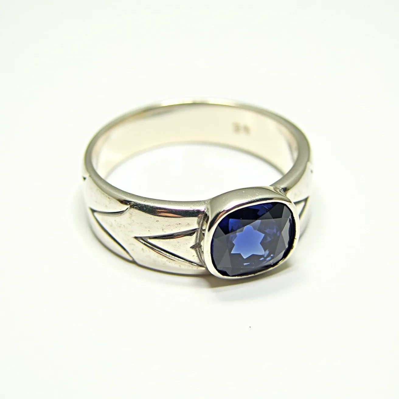 This men's silver ring features a striking blue gemstone at its center, elegantly set in a bezel setting. The gemstone is cushion-cut, giving it a modern yet timeless appeal. The band itself is crafted from silver and showcases engraved geometric patterns that add a touch of intricate detail. The design of the ring allows the stone to stand out prominently, making it a distinguished piece suitable for various occasions.