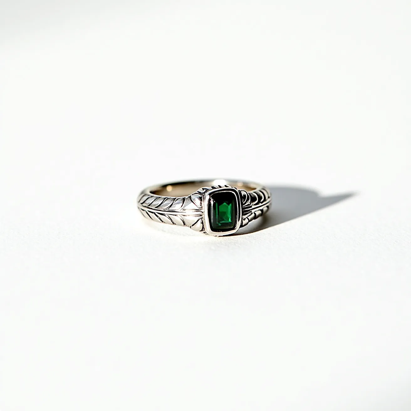 This men's silver ring features a striking design with a detailed band that includes engraved leaf patterns. At its center, there is a rectangular green gemstone, possibly an emerald or a green onyx, set securely in a bezel setting. The polished silver finish of the ring complements the deep green of the stone, creating a sophisticated contrast. The ring's construction lacks any additional clasps or attachments, focusing the design on the intricate detailing and the central gemstone.