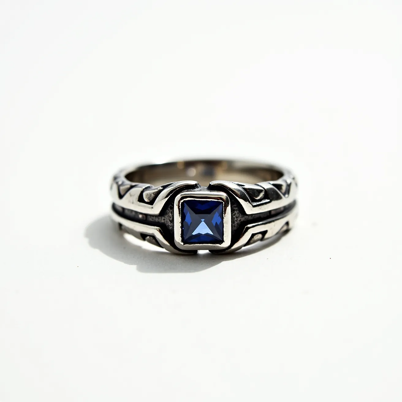 This men's silver ring features a square-cut blue stone set in a bezel setting, which provides a secure and stylish position for the gem. The band is intricately designed with geometric patterns that add depth and character to the overall appearance. Made primarily of silver, the ring exhibits a polished finish that complements the central stone's vibrant color. The absence of additional attachments or clasps highlights the ring's straightforward elegance, making it a distinctive accessory for its wearer.