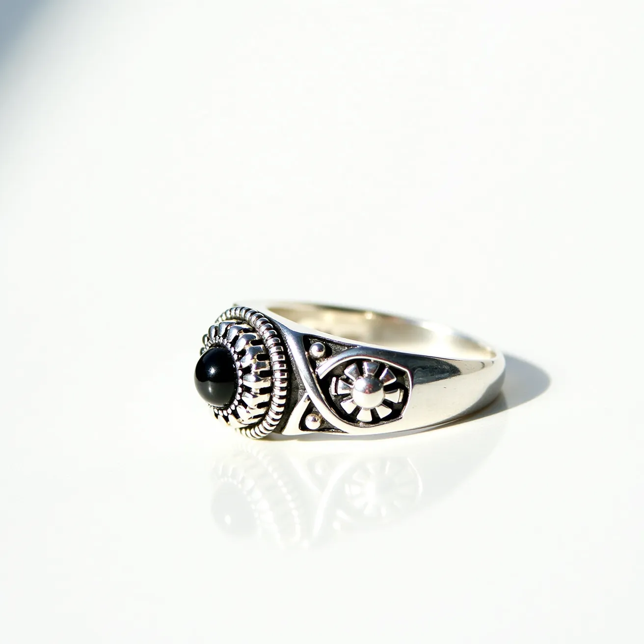 This men's silver ring features an intricate design with a central black stone. The stone is cut in a smooth, rounded shape and is set in a detailed, circular bezel that features a pattern of concentric lines. The sides of the ring display additional decorative elements, including a smaller raised circular motif with geometric patterns. The craftsmanship of the silver highlights the artistic detailing, creating a bold yet elegant accessory. The ring does not include any clasps or attachments, showcasing a solid band design.