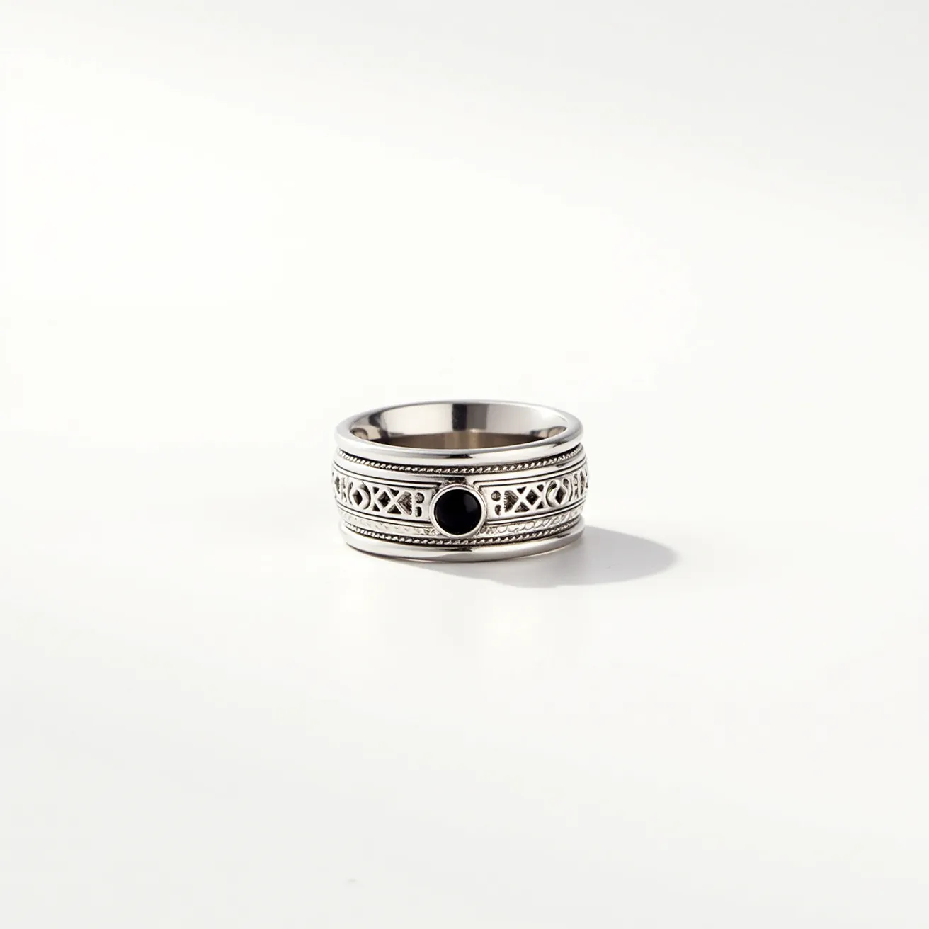 This men's spinner ring is crafted from a lustrous silver-toned metal, featuring an intricate design with repeating geometric patterns. At the center of the ring is a round, black stone, polished and set in a bezel style, providing a sleek contrast to the detailed band. The ring's spinner functionality allows the outer band to rotate independently around the inner ring, enhancing its tactile appeal and providing a dynamic accessory option. The combination of the richly detailed metalwork and the understated gemstone gives this piece a sophisticated yet modern look.