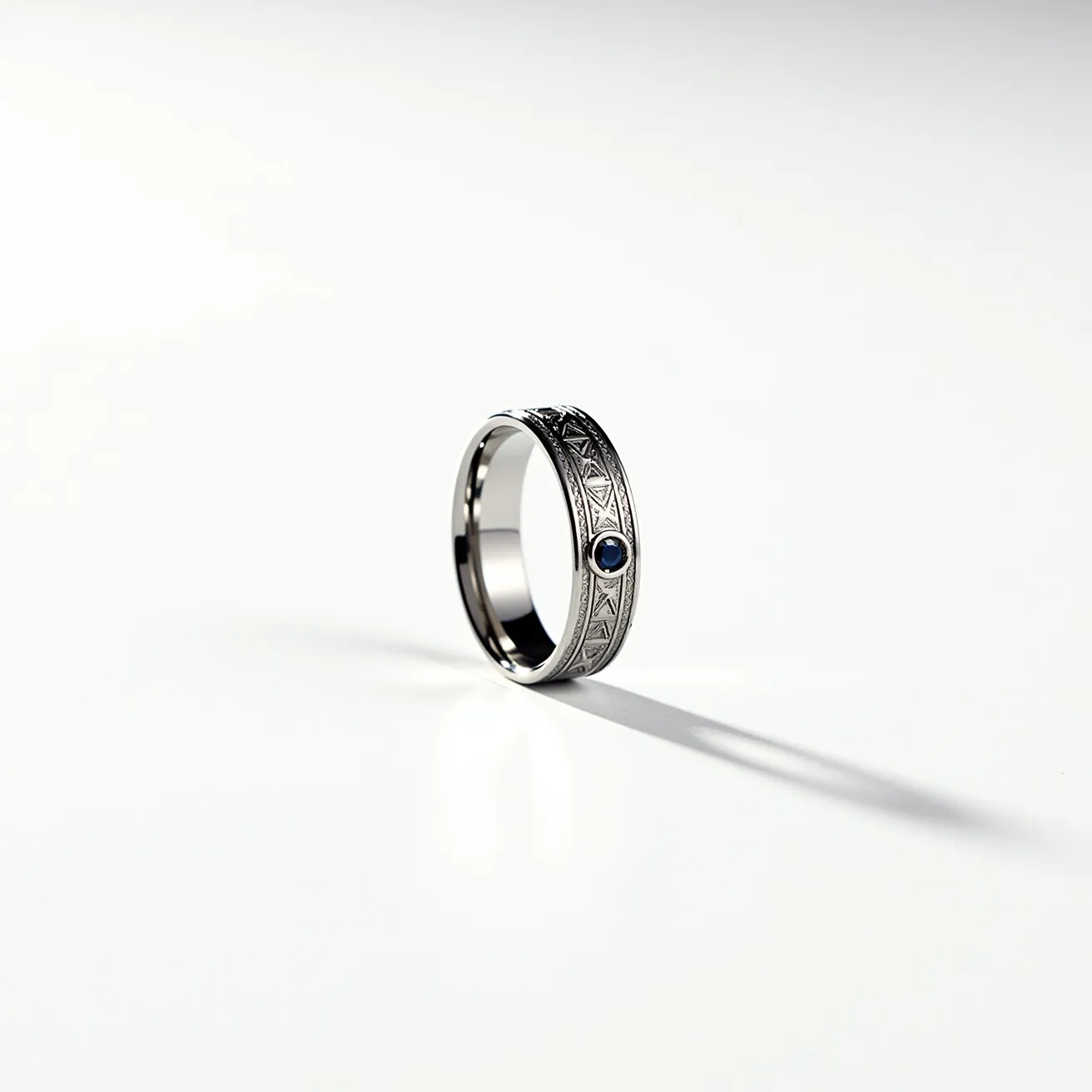 This men's wedding band is crafted from a polished metal, likely white gold or platinum, featuring an intricate engraved pattern along its surface. Set within the band is a single, round-cut blue sapphire, secured in a bezel setting that adds a touch of elegance and color contrast to the design. The combination of the metallic sheen and the deep blue gemstone creates a sophisticated and timeless look. The ring's design does not incorporate any clasps or attachments, showcasing a seamless circular form.