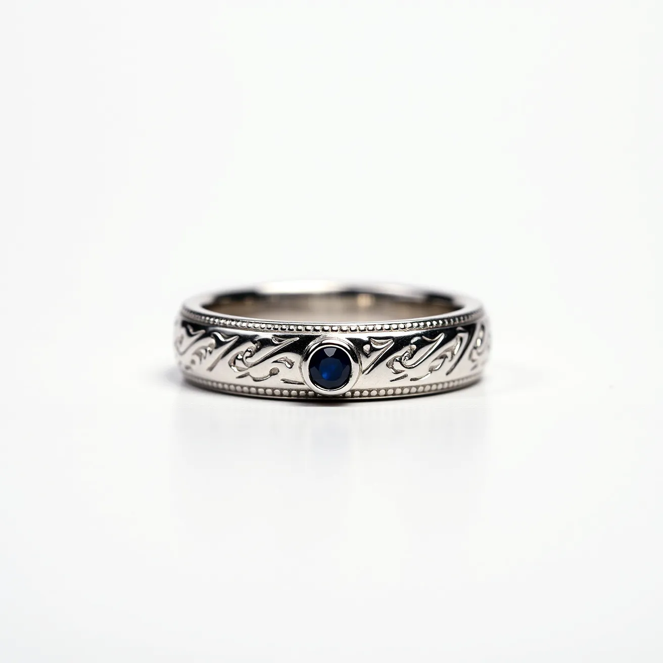 This men's wedding band features intricate detailing along its surface, crafted from what appears to be a polished, silver-toned metal. At the center is a round blue gemstone, which is securely held in place by a bezel setting, adding a touch of elegance and color contrast to the design. The band showcases an engraved pattern that weaves across its circumference, enhancing its decorative appeal. The careful craftsmanship is evident in the symmetrical design and smooth finish, resulting in a timeless piece that is both stylish and sophisticated.