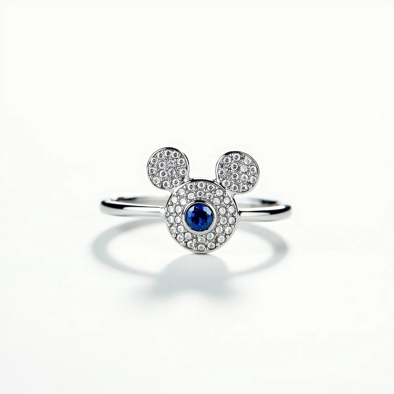 This Mickey Mouse ring features a polished metal band likely made from a platinum or white gold alloy, complementing its elegant design. The iconic Mickey Mouse head is adorned with a pavé setting of small, sparkling diamonds or diamond-like stones, creating a glamorous texture. At its center, a larger, round-cut blue sapphire or sapphire-like stone is bezel-set, adding a striking contrast and focal point to the piece. The ring is crafted with seamless precision, showcasing a whimsical yet refined aesthetic without any visible clasps or attachments, ensuring a continuous and elegant finish.