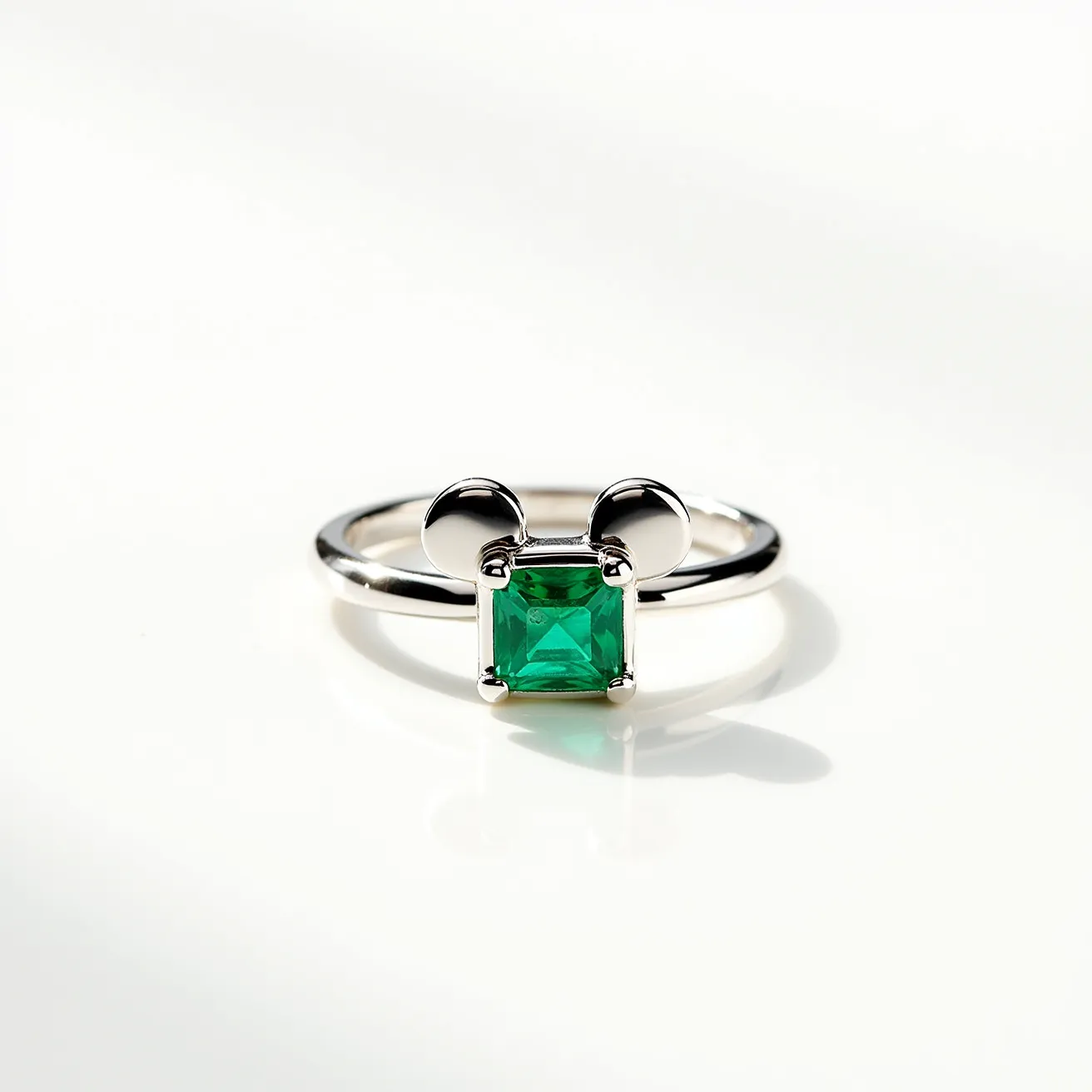 This Mickey Mouse ring features a sleek metal band crafted possibly from sterling silver or white gold, giving it a polished appearance. The centerpiece is a vibrant, square-cut green gemstone, securely held in place by a four-prong setting. Above the gemstone, two rounded metallic elements resemble Mickey Mouse ears, adding a whimsical touch to the design. The ring does not appear to have any visible clasps or attachments, maintaining a simple yet elegant structure.