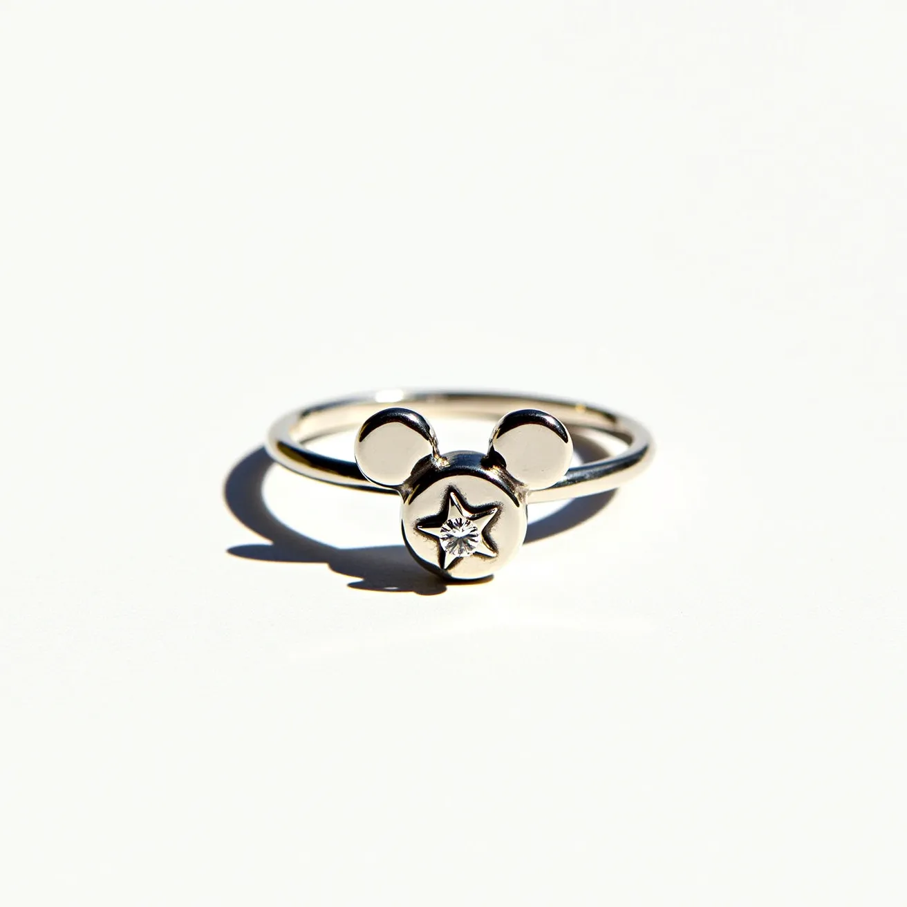 This Mickey Mouse ring features a sleek, minimalist design crafted from a polished silver material that gives it an elegant shine. The iconic Mickey Mouse head motif serves as the centerpiece, adorned with a small, centrally placed gem. The gem is a clear stone, possibly a diamond or a cubic zirconia, cut into a star shape and set flush within the circular face of the motif, highlighting its intricate design. The band is simple and rounded, complementing the overall understated yet playful aesthetic of the ring.