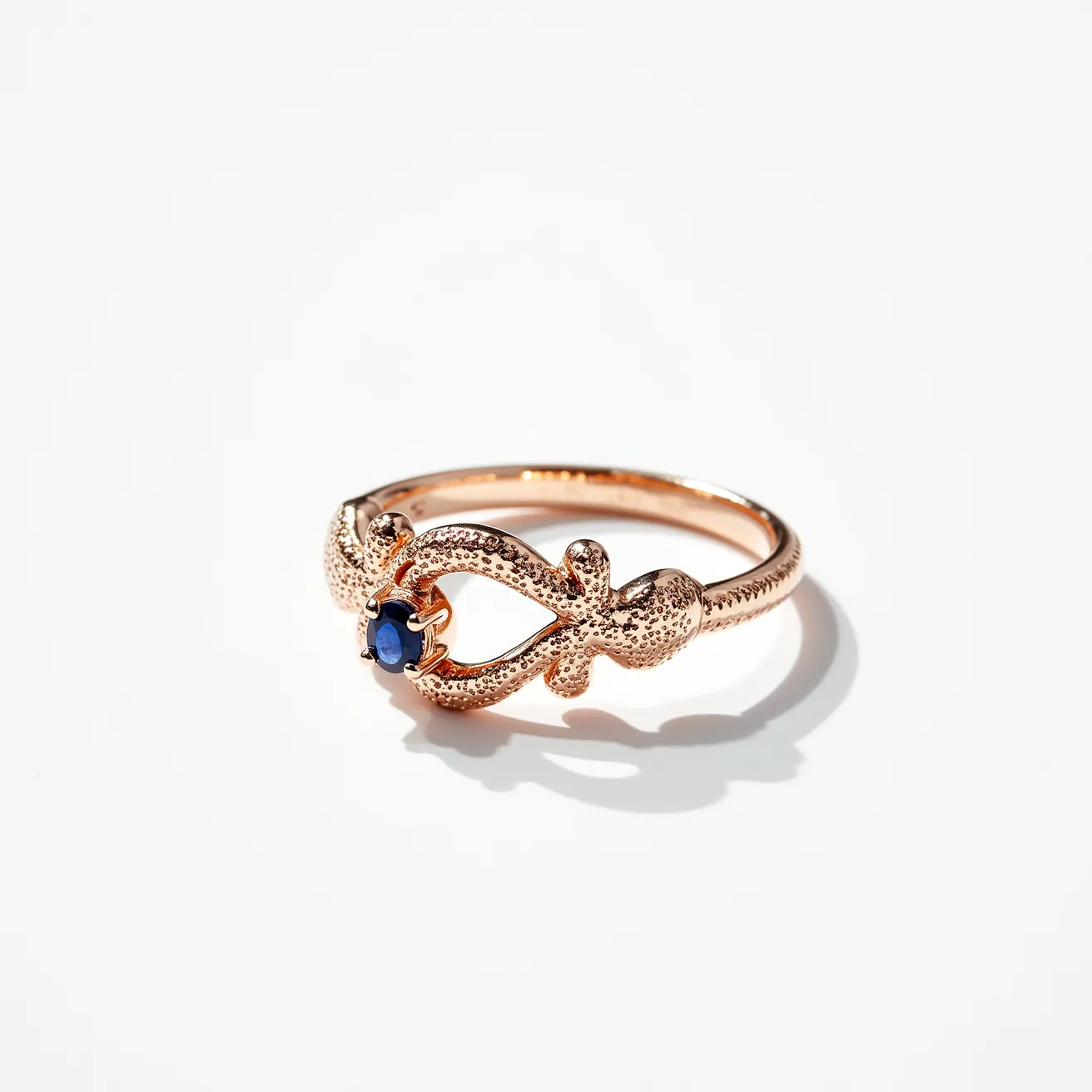 This midi ring features a delicate, ornate design crafted from a warm, rose gold-toned metal, with intricate texturing adding an antique feel. At its center, it showcases a small, round-cut deep blue gemstone, likely a sapphire, securely set in a classic four-prong setting, enhancing its elegant appeal. The ring's structure follows a smooth, seamless band without any visible clasps or attachments, emphasizing its simplicity and ease of wear.