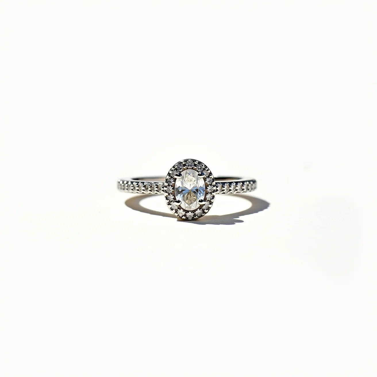 This moissanite promise ring features an oval-cut moissanite gemstone at its center, surrounded by a halo of smaller, round stones that enhance its brilliance. The band appears to be made of a lustrous metal, likely white gold or platinum, as it complements the sparkling quality of the stones. The band itself is adorned with a row of tiny, shimmering stones that continue the theme of elegance and sophistication. The setting and arrangement provide a sense of harmony and grace, making it an exquisite token of commitment.