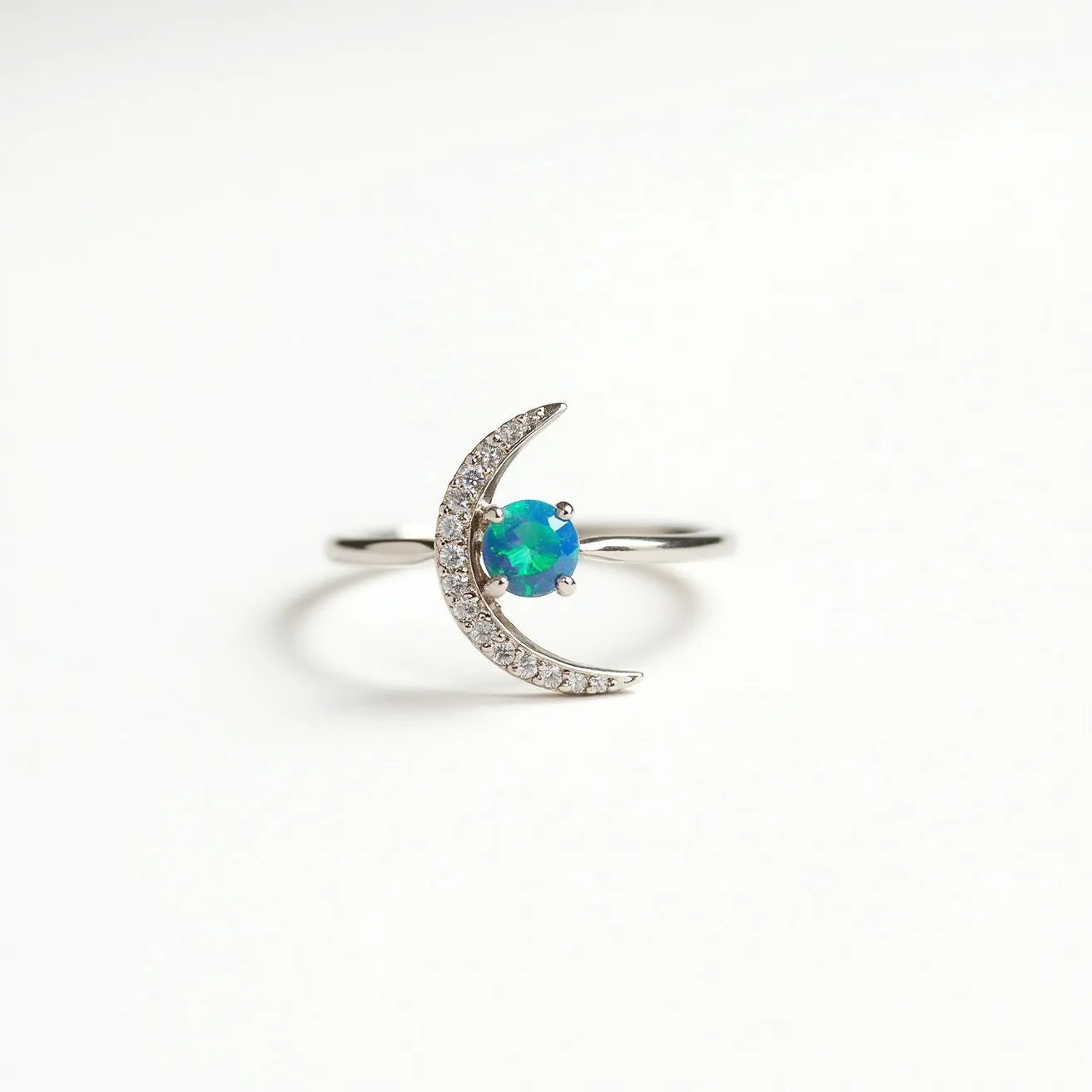 This moon ring features a delicate crescent crafted from what appears to be a high-quality metal, possibly sterling silver or white gold, gracefully wrapping around a central, round opal gemstone. The opal, exhibiting vibrant blue and green hues, is securely held in place by a pronged setting, allowing maximum visibility and light play. The crescent is embellished with a series of small, sparkling clear stones—likely diamonds or cubic zirconia—set in a pavé style, adding a shimmering contrast to the colorful opal centerpiece. The band itself is slender and polished, enhancing the overall elegance of the piece.