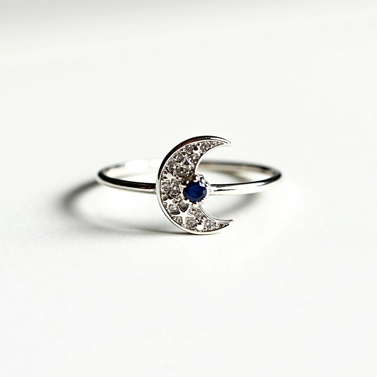 This moon ring is elegantly crafted from a shiny silver material, forming a delicate band. The centerpiece features a crescent moon design, studded with small, sparkling clear gemstones that appear to be set in a pavé setting. At the center of the crescent moon is a striking round-cut blue gemstone, securely held in place by a prong setting, adding a touch of color and contrast to the piece. The overall design is minimalist yet enchanting, highlighting the celestial theme with a modern finish.