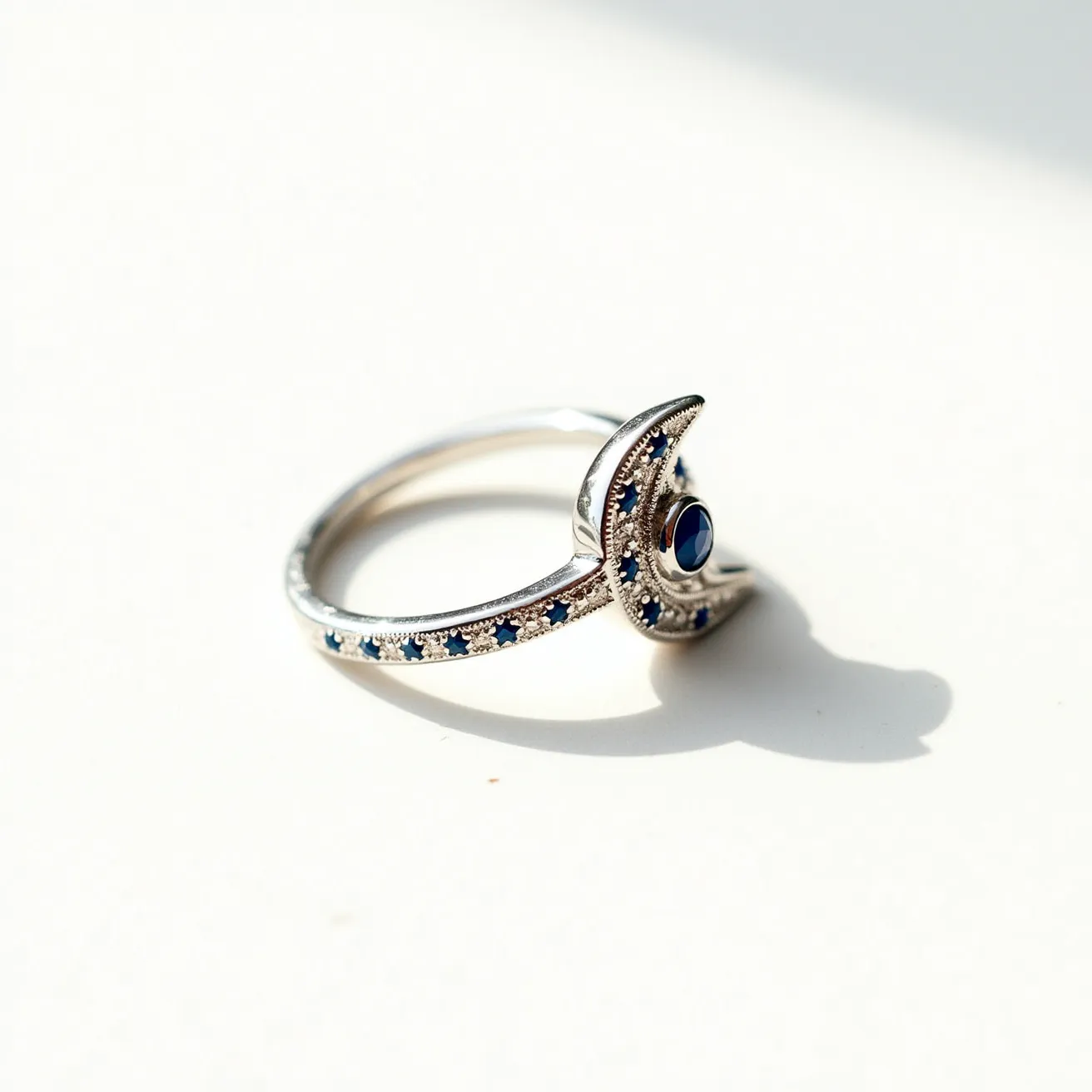 This moon ring features a crescent shape made of a silvery metal, likely sterling silver, which gives it a sleek and modern appearance. The crescent is adorned with small, round-cut blue gemstones, possibly sapphires, that are set along its curve, providing a striking contrast against the metal. At the center of the crescent is a larger, round blue stone set securely within a bezel setting. The elegant arrangement of the stones highlights the crescent design, and the sleek band seamlessly integrates with the overall craftsmanship of the ring.