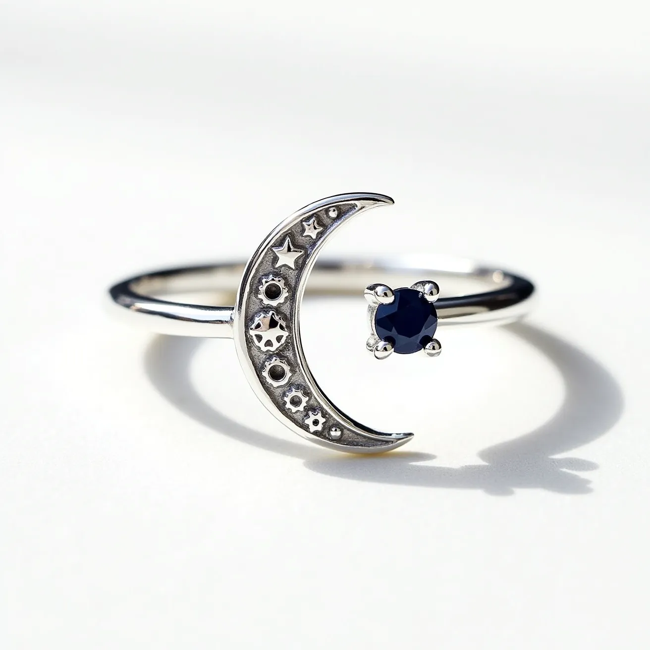 This moon ring features a sleek silver band with a crescent moon design on one side, subtly embellished with star-shaped details. Adjacent to the crescent is a dark blue gemstone, likely a sapphire, cut in a round shape and secured in a prong setting. The prongs delicately hold the gem, enhancing its rich color and brilliance, while the minimalist band allows the celestial themes and gemstone to stand out prominently.