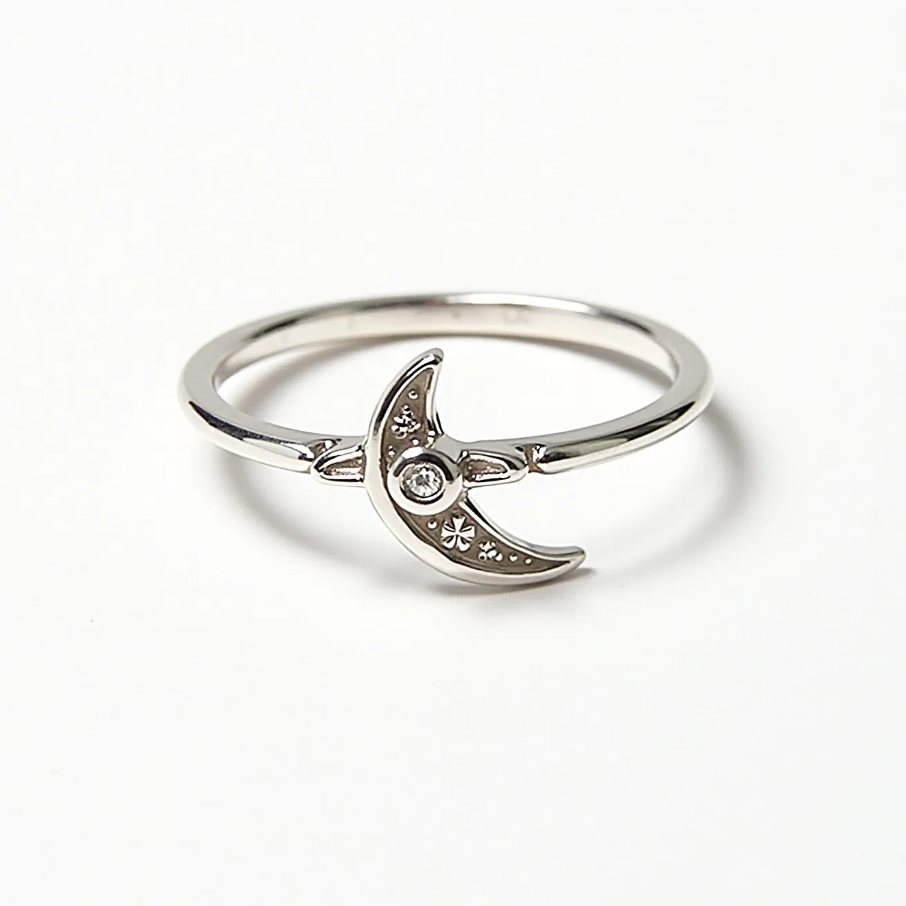 This moon ring features a crescent moon design crafted from what appears to be a polished metal, likely silver or white gold. At the center of the crescent is a small, round-cut gemstone, possibly a diamond or a similar clear stone, set in a bezel setting that securely holds the gem in place. The crescent itself is adorned with delicate star-like engravings, adding an element of celestial charm to the ring. The band is smooth and unadorned, complementing the understated elegance of the moon motif.