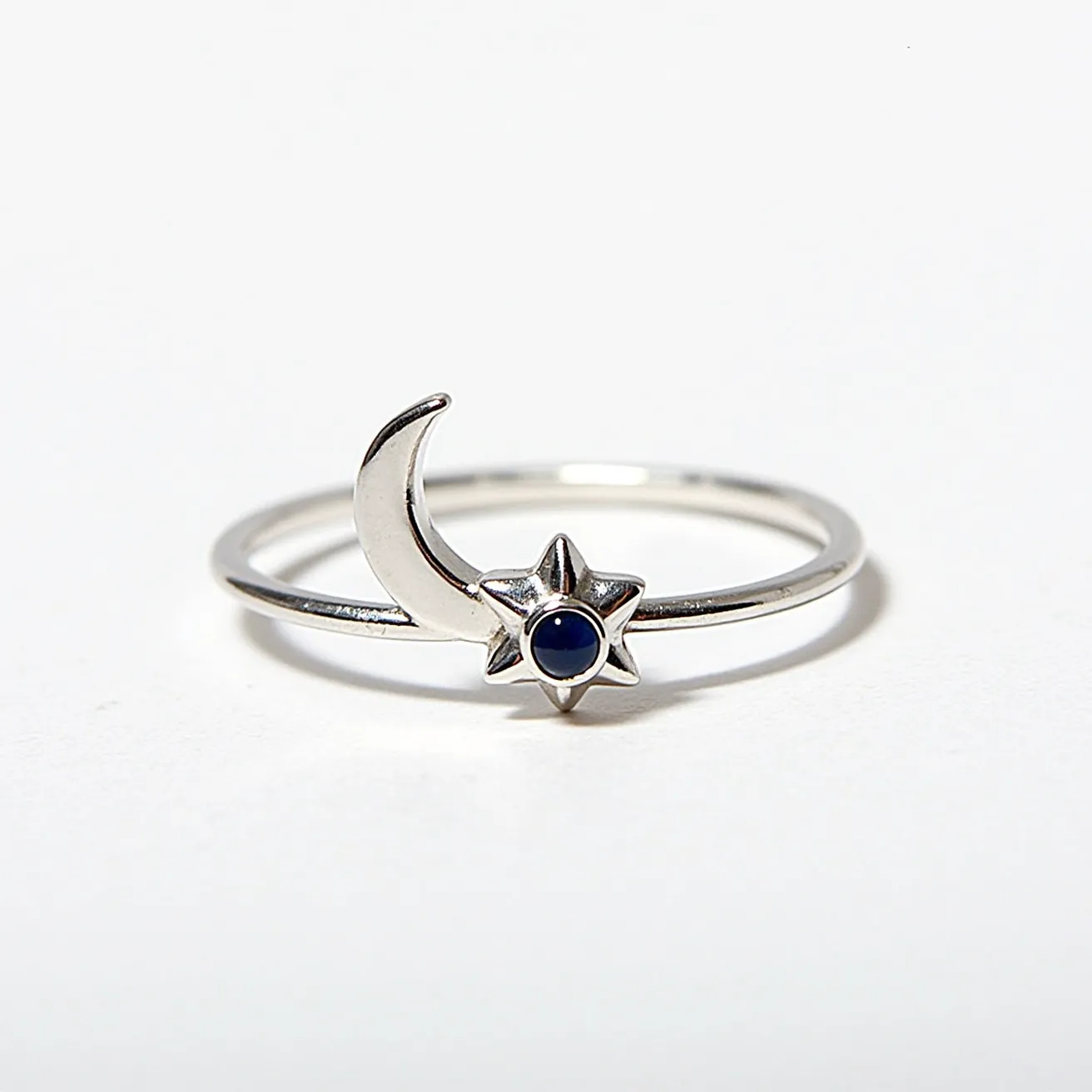 This moon ring features a slender band crafted from a silvery metal, most likely sterling silver, lending it a sleek and elegant appearance. It prominently displays a crescent moon design next to a charming star motif. The star is adorned with a small, round, deep blue gemstone, likely a sapphire or a similar stone, held securely in a bezel setting which integrates seamlessly with the star's design. The overall composition is minimalist and celestial, creating a piece that captures the night sky's enchanting allure.