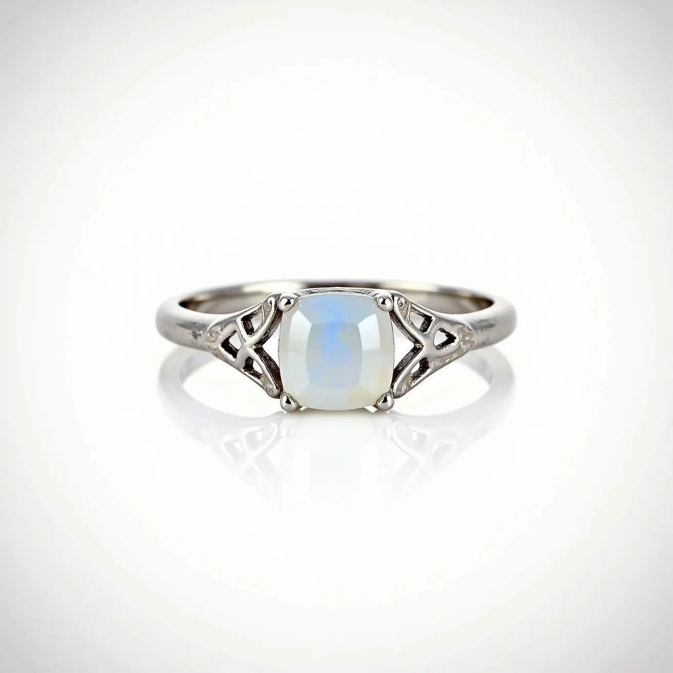 This moon ring features a delicate design crafted from a polished metal, likely silver, which provides a sleek and elegant base. The centerpiece of the ring is a cushion-cut moonstone, showcasing its characteristic milky sheen with subtle flashes of blue. The gem is held securely in place by a four-prong setting, adding both protection and an element of classic style. Flanking the center stone are intricately designed openwork details, adding a touch of vintage charm to the overall aesthetic. The band is slim and smooth, contributing to the ring's refined simplicity and allowing the moonstone to take center stage.