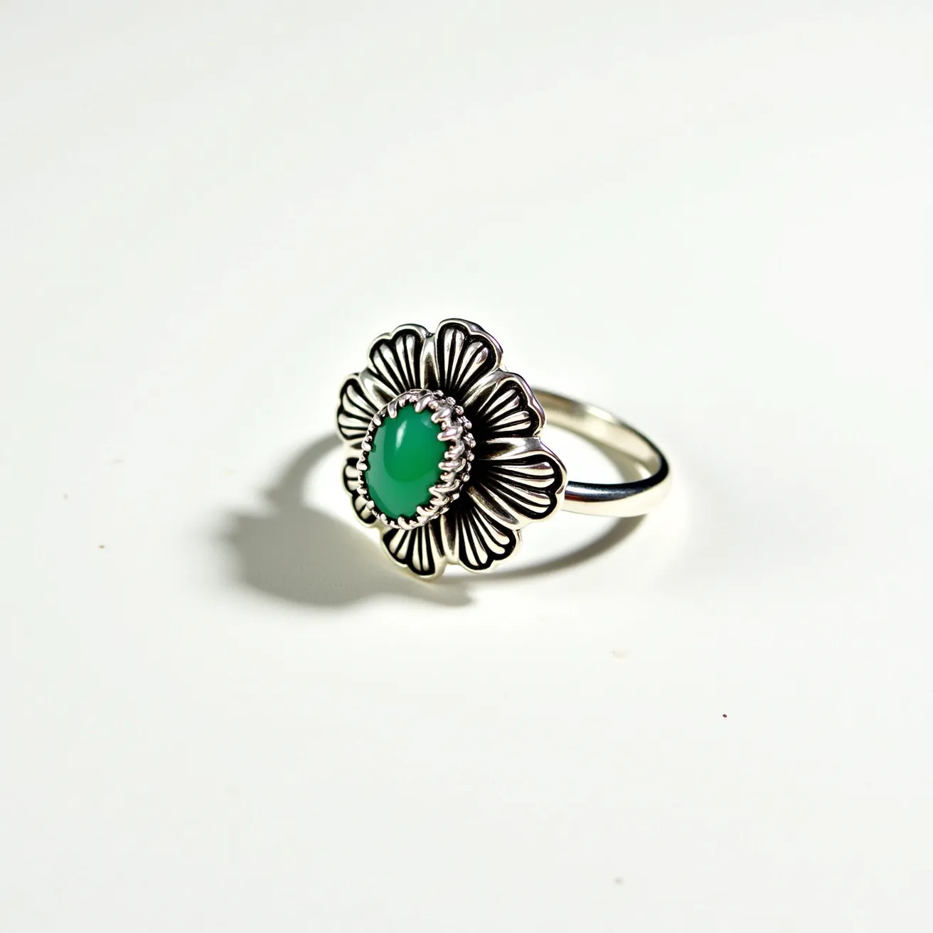 This moss agate ring features a striking green oval cabochon stone set at its center, surrounded by a scalloped bezel setting that resembles the petals of a flower. The intricate metalwork forming the flower petals appears to be crafted from a polished silver material, giving it a detailed and elegant look. The band of the ring is a simple, smooth silver, allowing the decorative floral feature to stand out. This design effectively highlights the natural beauty of the moss agate while incorporating an artistic floral motif.