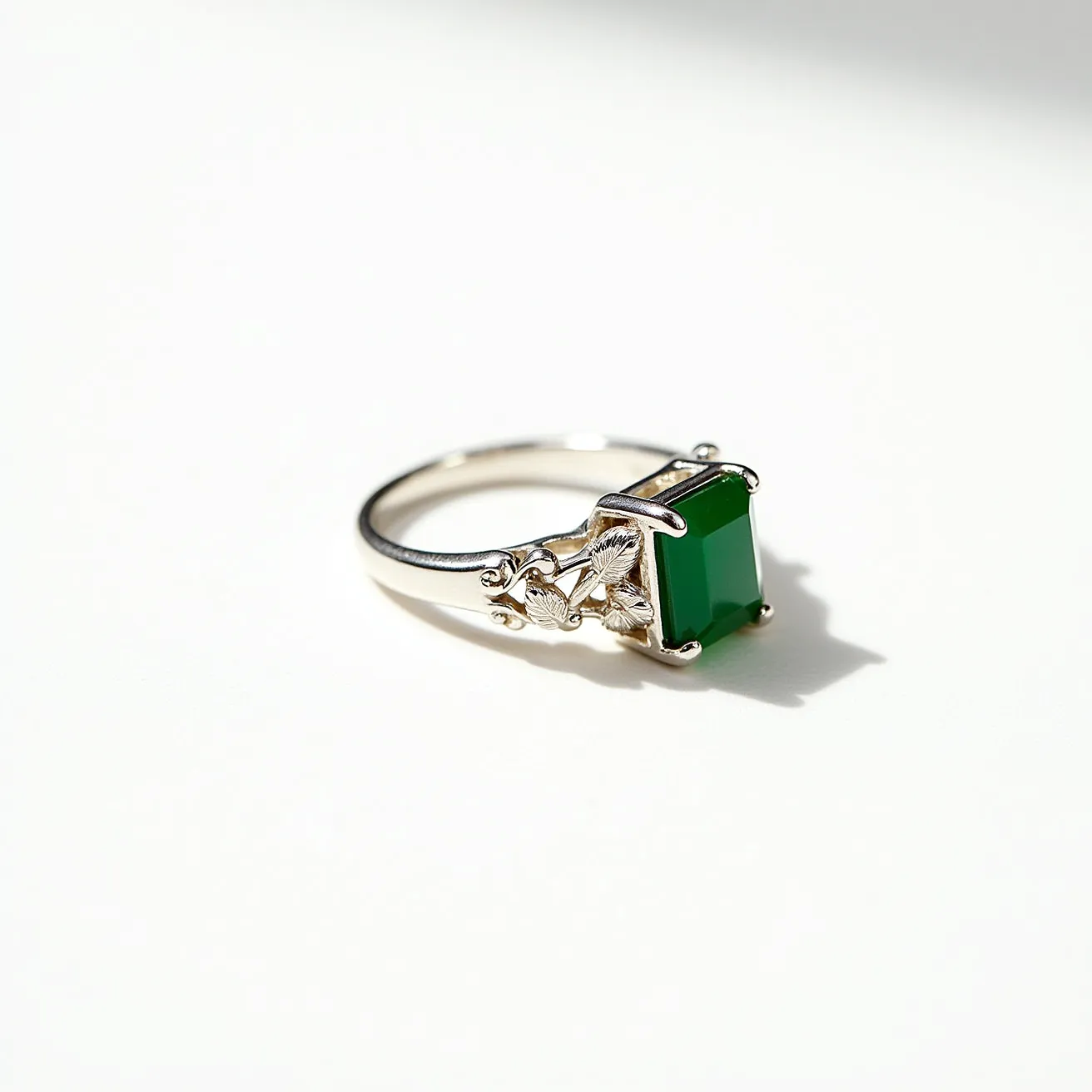 This moss agate ring features a vibrant, square-cut green stone set in a silver band. The setting is intricate, with decorative floral or leaf motifs on the band, enhancing its elegant design. The stone is securely held in place by prongs at each corner. The band appears smooth and polished, complementing the overall aesthetic of the ring.