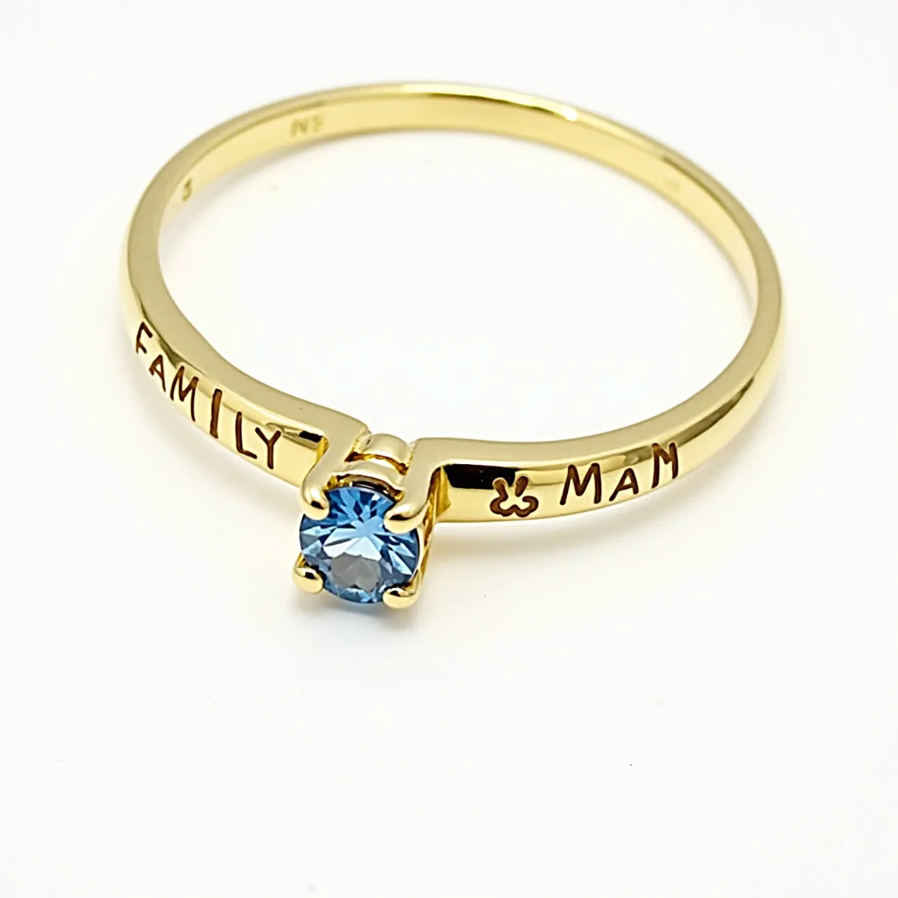 This mother's ring features a sleek, gold band inscribed with the words "FAMILY" and "MAM." At its center, the ring showcases a single round-cut blue gemstone, held securely in place by a prong setting that highlights its vibrant color and sparkle. The elegant design and thoughtful engravings make this ring a meaningful piece of jewelry.