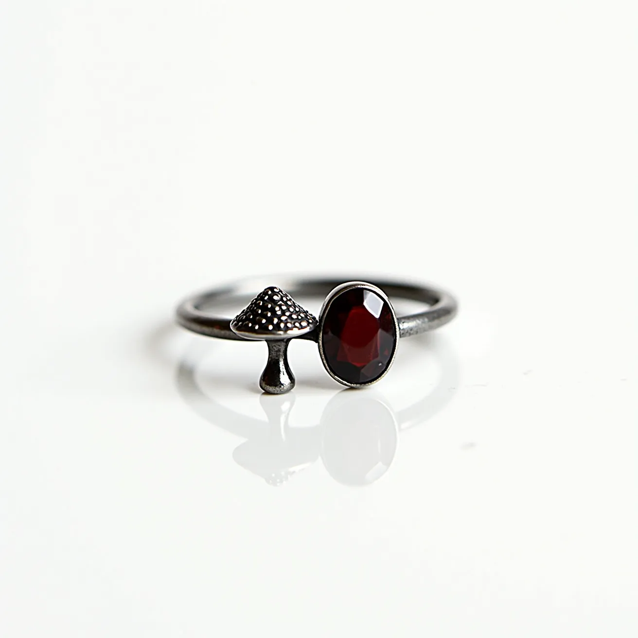 This mushroom ring features a metallic band adorned with a whimsical mushroom motif, showcasing a textured cap atop a slender stem. Adjacent to the mushroom is an oval-cut gem with a deep red hue, securely set in a bezel setting, which enhances the stone's rich color and adds a touch of elegance to the design. The minimalist design combines nature-inspired elements with a modern aesthetic, making it a unique accessory.