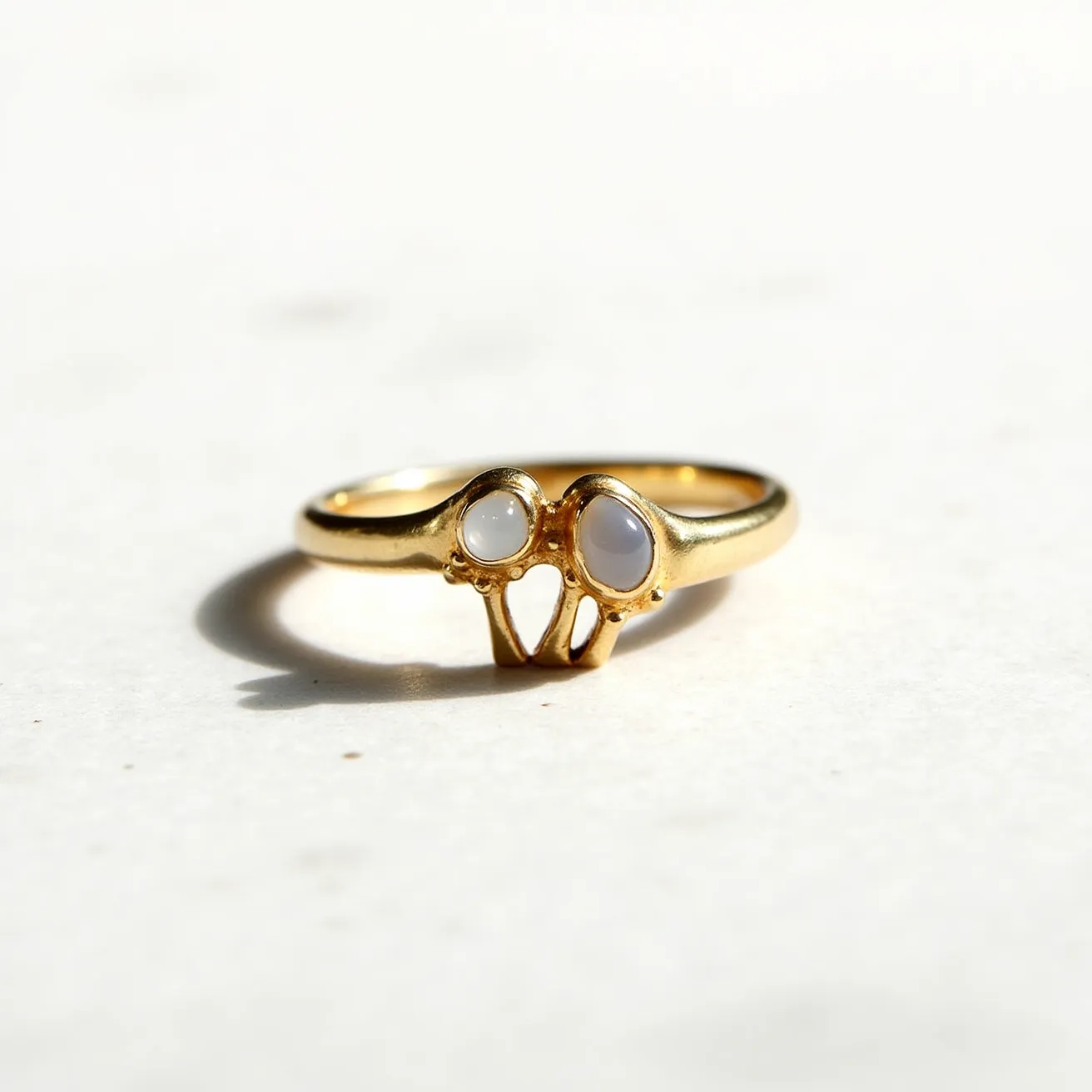 This mushroom ring features a gold band crafted to resemble two mushroom shapes. Each cap of the mushrooms is adorned with a stone that appears to be a cabochon-cut opal, exhibiting a translucent and lustrous quality. The stones are set into the gold in a bezel setting, which secures them smoothly within the design without any prongs. The band itself is sleek and rounded, providing a consistent and polished appearance. There are no visible clasps or attachments, indicating that the ring has a classic circular design meant for easy wearing.