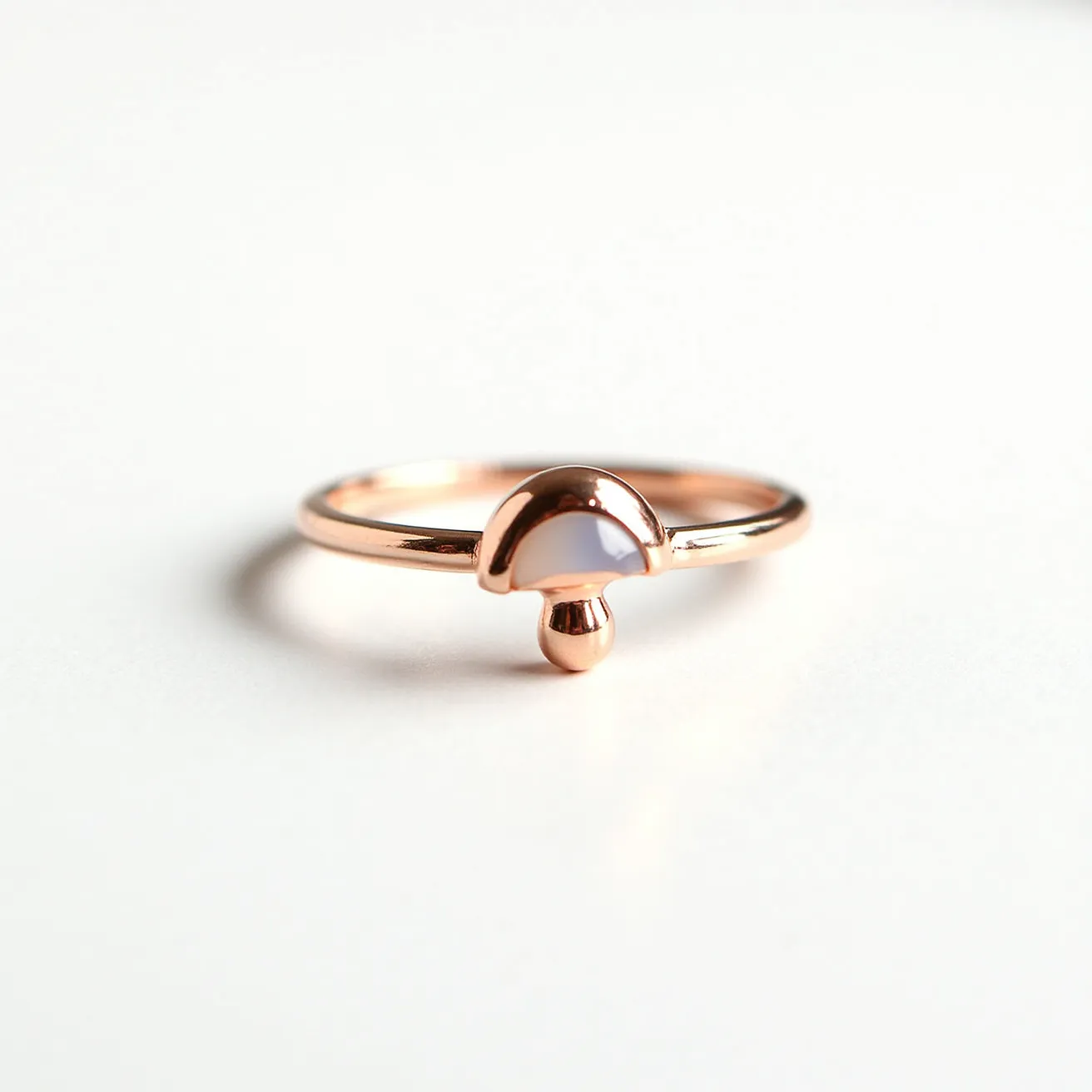 This mushroom ring features a minimalist design crafted from a polished rose gold metal, embodying a delicate and elegant aesthetic. The focal point of the ring is a mushroom-shaped motif that prominently displays a single, smooth cabochon gem set flush into the cap, likely an opalescent moonstone that adds a subtle touch of color and luminescence. The stone's shape complements the curved lines of the mushroom structure, seamlessly integrated into the shank. The gleaming finish of the metal and the stone together create a unified and whimsical piece, showcasing careful craftsmanship without the presence of any visible clasps or attachments, as is typical with rings of this nature.