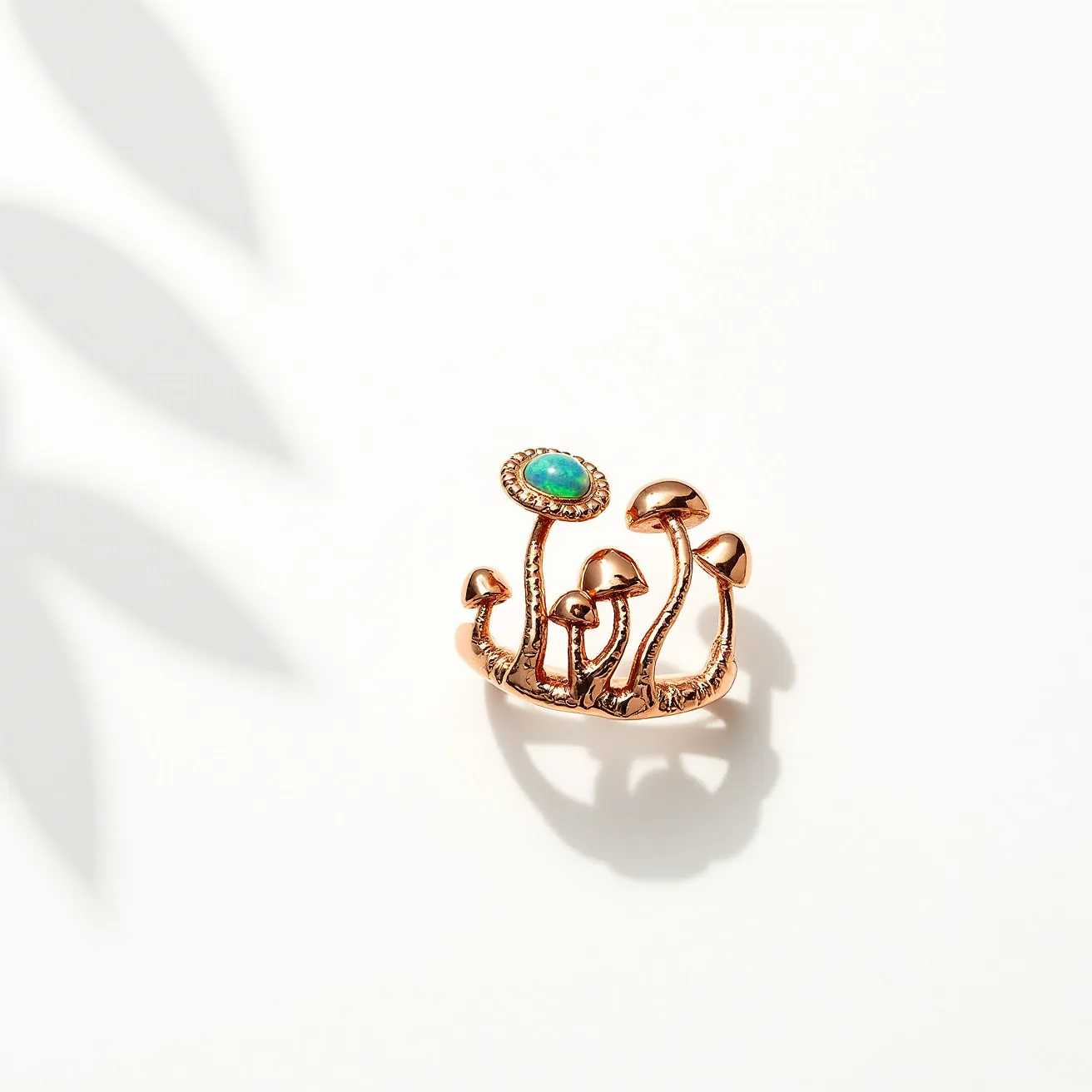 This mushroom ring is crafted from a coppery metal, featuring a whimsical design of slender mushroom stems. A standout element is the bright, round gemstone crowning one of the taller mushrooms, which resembles a turquoise cabochon with its distinct blue-green hue. The gem is set in a bezel setting, secured with a decorative border that complements the organic theme. The ring’s band is unified in design with the mushrooms, creating a cohesive and artistic piece without visible clasps or additional attachments, emphasizing its seamless nature.