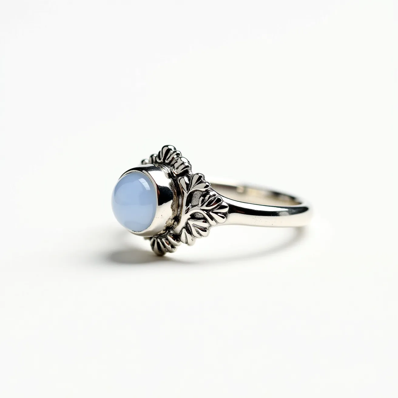 This mushroom ring features a silver band adorned with detailed leaf-like designs surrounding the central gem. The gemstone is a smooth, round cabochon cut, with a subtle, translucent blue hue, reminiscent of chalcedony. Its setting is a bezel, providing a secure and seamless transition between the stone and the band. This elegant combination of materials and design creates a whimsical and nature-inspired aesthetic.