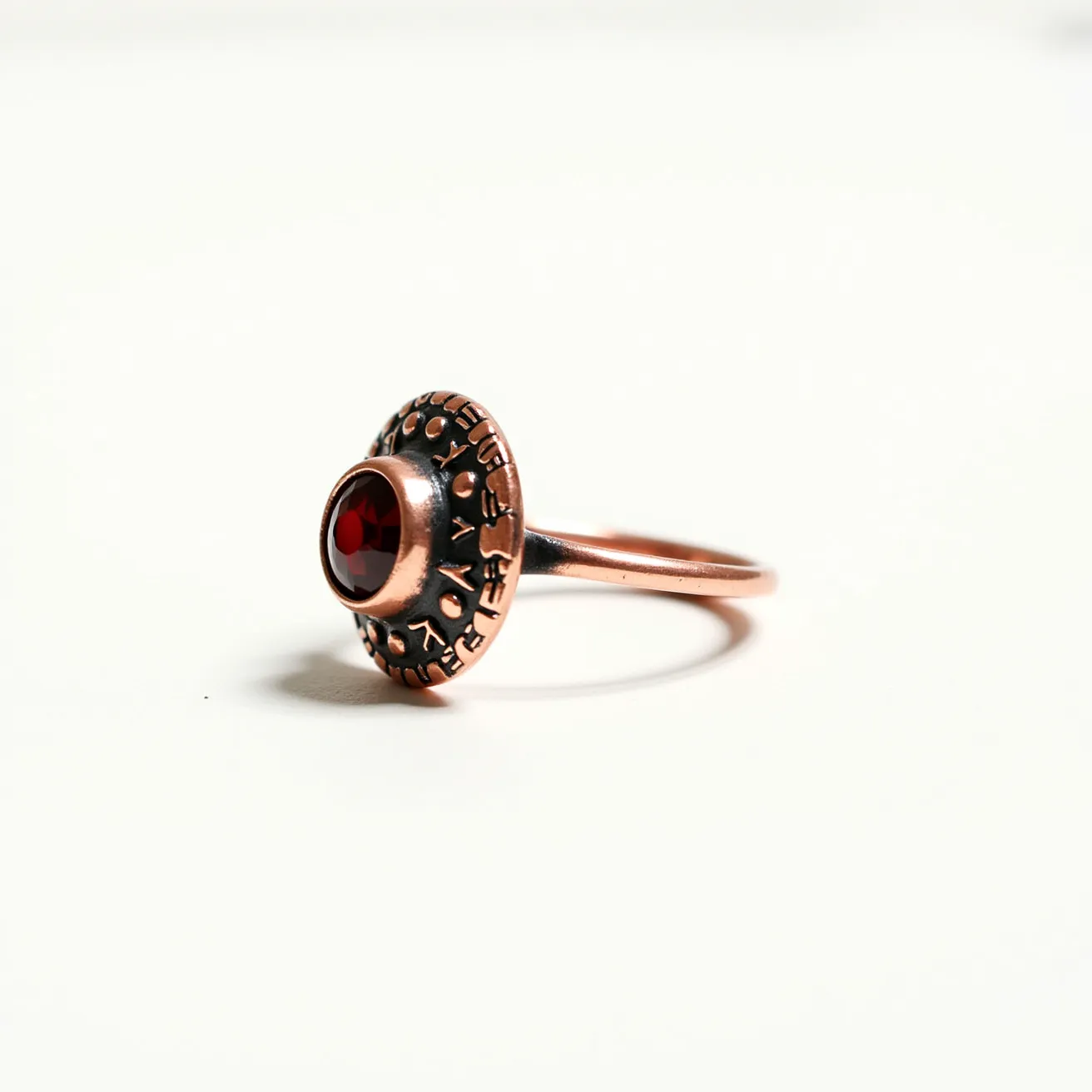 This mushroom ring features a unique design crafted from a rosy metallic material, possibly copper or rose gold. The standout feature is a round, deep red gem set at the center, likely cut in a cabochon style to enhance its rich hue. The gem is bezel-set, securely encased within the metal, which adds a vintage allure to the piece. The shank of the ring is smooth and slender, complementing the intricately designed, textured mushroom cap adorned with engraved or embossed patterns, enhancing the overall whimsical aesthetic.