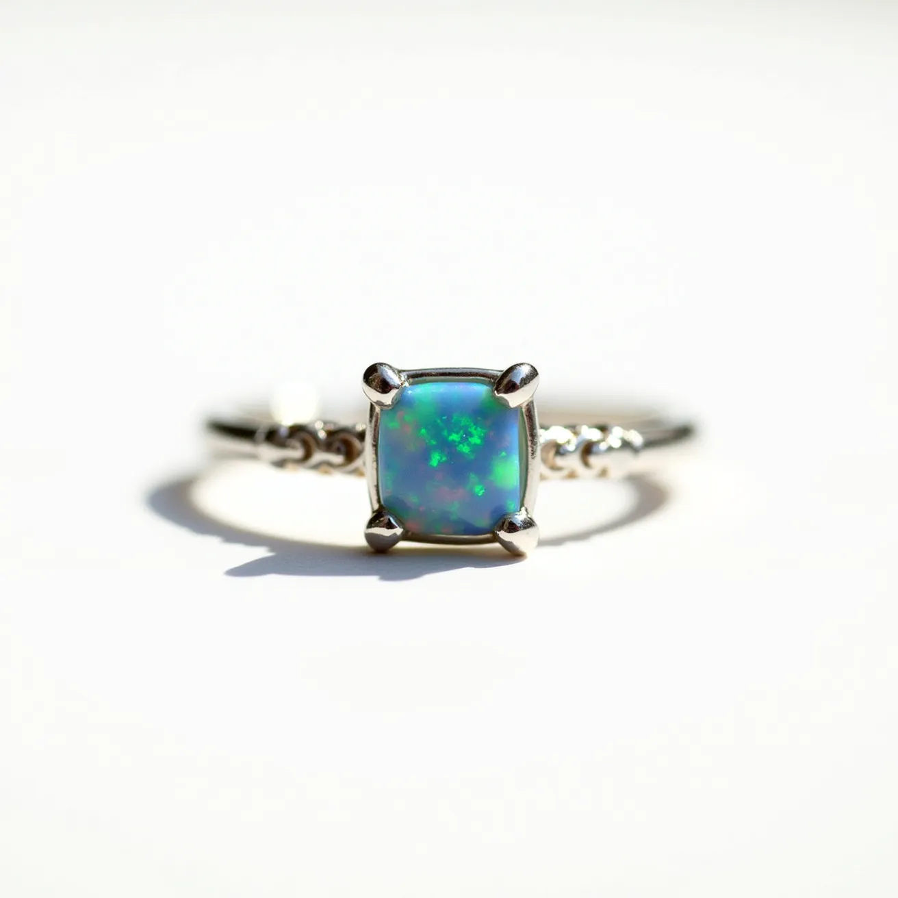 This October birthstone ring features a stunning opal as its centerpiece, showcasing a mix of blue and green hues that capture the light beautifully. The gem is cut in a cushion shape, enhancing its natural play of color, and is set in a four-prong setting that secures it elegantly. The band appears to be crafted from a polished metal, likely silver or white gold, adding a complementary shine to the iridescent opal. Delicate detailing on the band adds a touch of sophistication to the overall design.