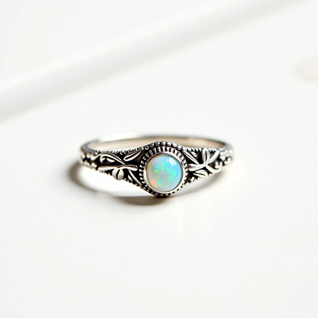 This October birthstone ring features a polished opal as the central gemstone, showcasing its characteristic play of colors with hints of blues and greens. The opal is cut into a smooth cabochon, enhancing its natural iridescence. The stone is securely set in a bezel setting, which is surrounded by intricate metalwork, likely made of sterling silver, adding a vintage or antique flair with its engraved floral details. The band of the ring is delicately crafted, complementing the opal and providing a harmonious blend of elegance and artistry.