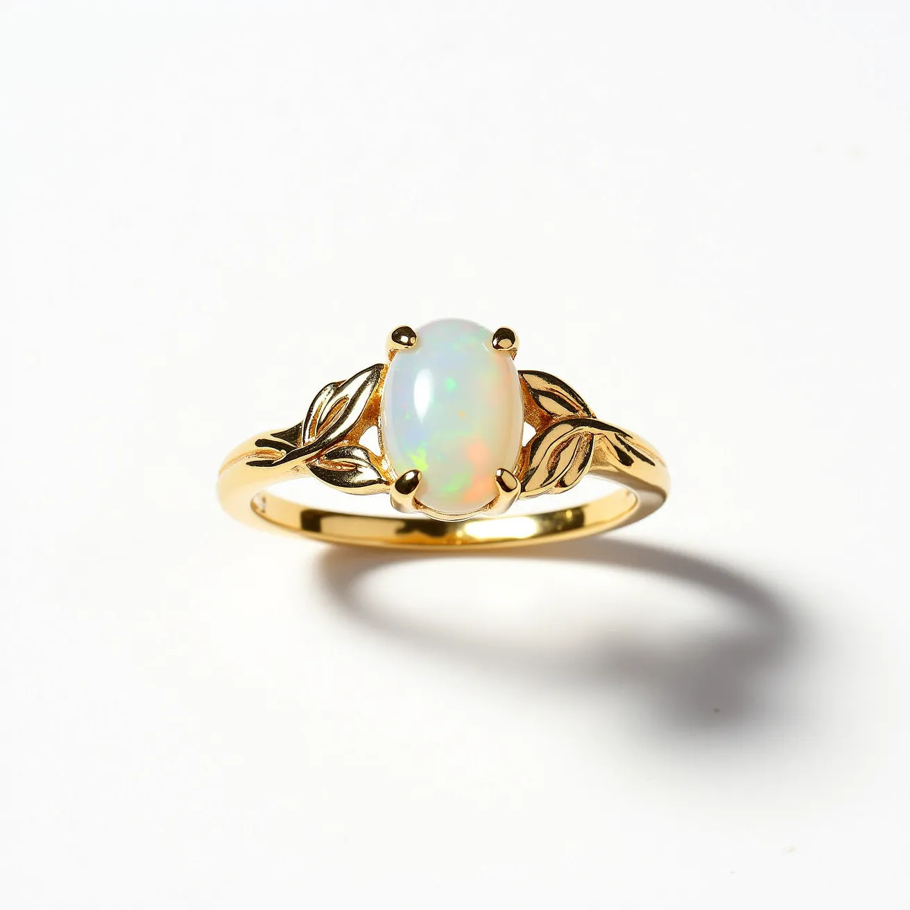 This October birthstone ring features a central opal gemstone, which is smooth and oval-cut. The opal, known for its iridescent play of color, is held securely in place by a classic prong setting. The band of the ring is made of gold and is elegantly designed with leaf motifs on either side of the stone, adding a touch of natural elegance to the piece. The overall composition of the ring highlights both the beauty of the opal and the intricate gold detailing.