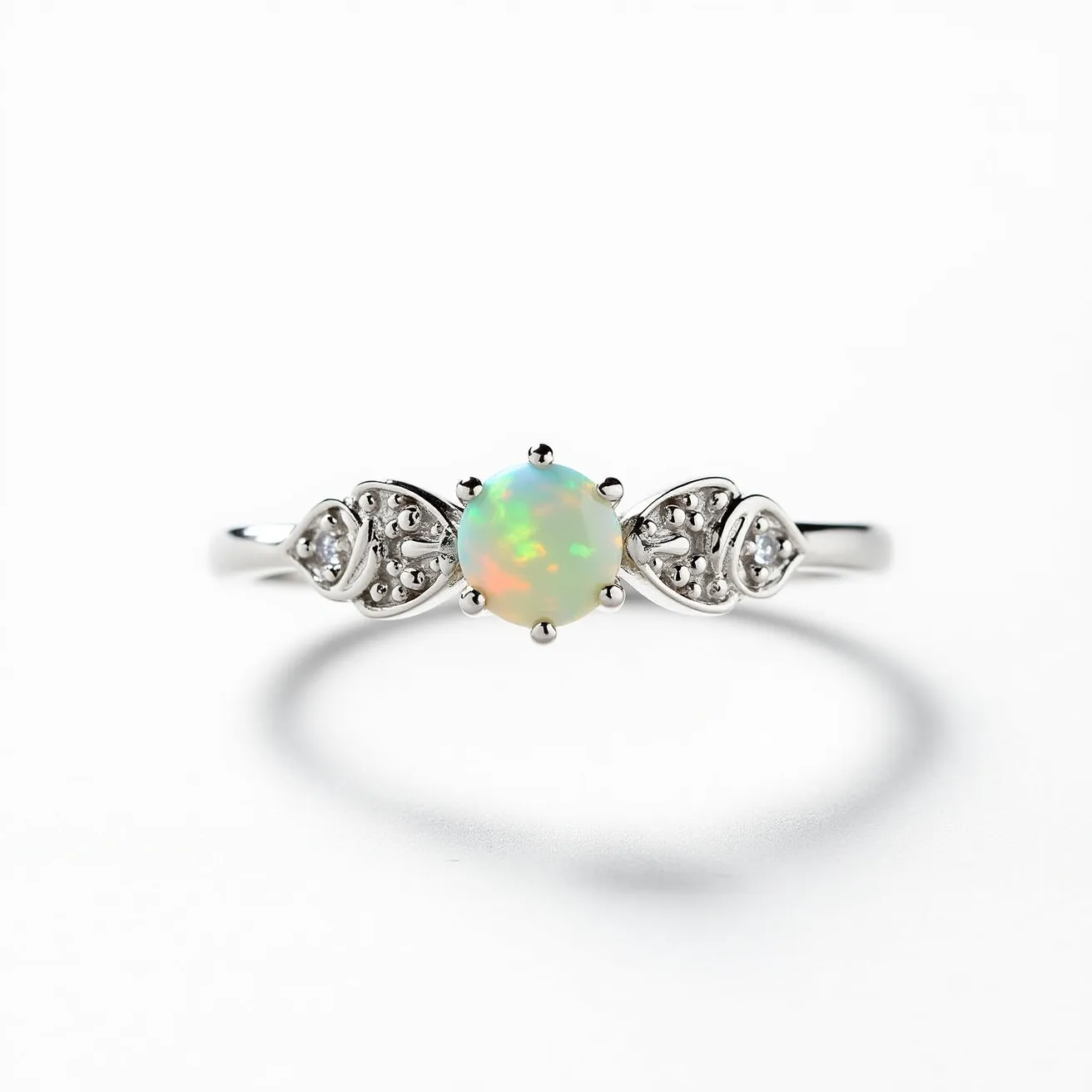 This opal engagement ring features a central round opal stone set in a classic prong setting. The opal exhibits a vivid play of colors, characteristic of its unique iridescent qualities. Flanking the opal are small, round accent diamonds set in a decorative, leaf-like pattern crafted in a white metal band, likely white gold or platinum, lending the ring a delicate and elegant appearance. The design is both modern and timeless, complementing the opal’s natural beauty.