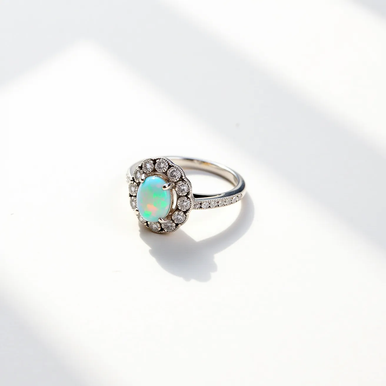 This opal engagement ring features an enchanting oval-shaped opal as its central gemstone, displaying a mesmerizing play of colors. The opal is encircled by a halo of round, brilliant-cut diamonds, providing a sparkling contrast to the central stone. The setting is crafted from a lustrous white metal, likely white gold or platinum, which enhances the brightness of the gemstones. The band is elegantly understated, possibly adorned with small pavé-set diamonds that add additional shimmer to the piece. The opal is secured with a classic four-prong setting, ensuring the stone is safely anchored while maximizing its exposure to light.