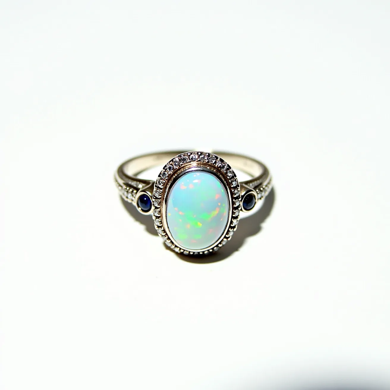 This opal ring features a central opal gem with an oval cut, showcasing a play of vivid colors. The opal is surrounded by a halo of small sparkling stones set in a detailed metallic band, possibly silver or white gold. Additionally, there are two small round blue stones set into the shank on either side of the opal, adding an elegant touch. The intricate settings enhance the overall design, creating a striking and sophisticated piece of jewelry.