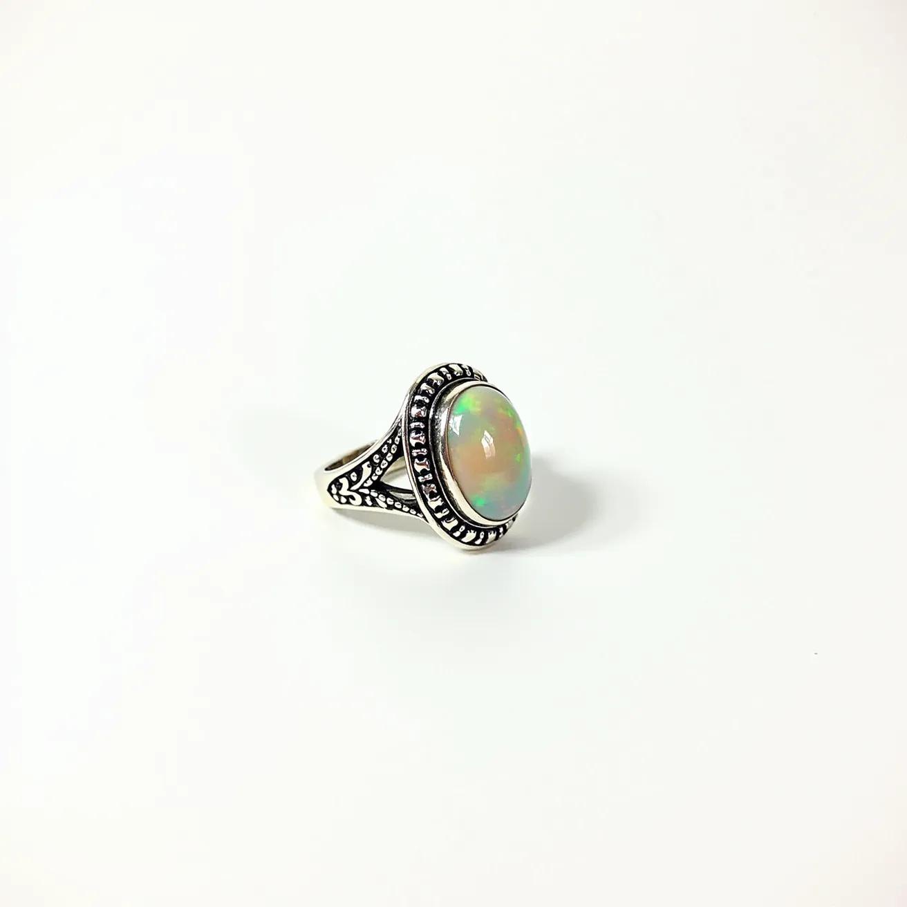 This opal ring features a captivating oval opal as its centerpiece, displaying a play of colors typical of high-quality opals. The opal is set in a bezel style, surrounded by an intricately patterned silver setting that accentuates the stone's natural beauty. The band is crafted from silver and showcases engraved detailing that adds an elegant and vintage feel to the overall design. The ring design emphasizes both the opal's vibrant hues and the craftsmanship of the silver setting, making it a striking piece of jewelry.