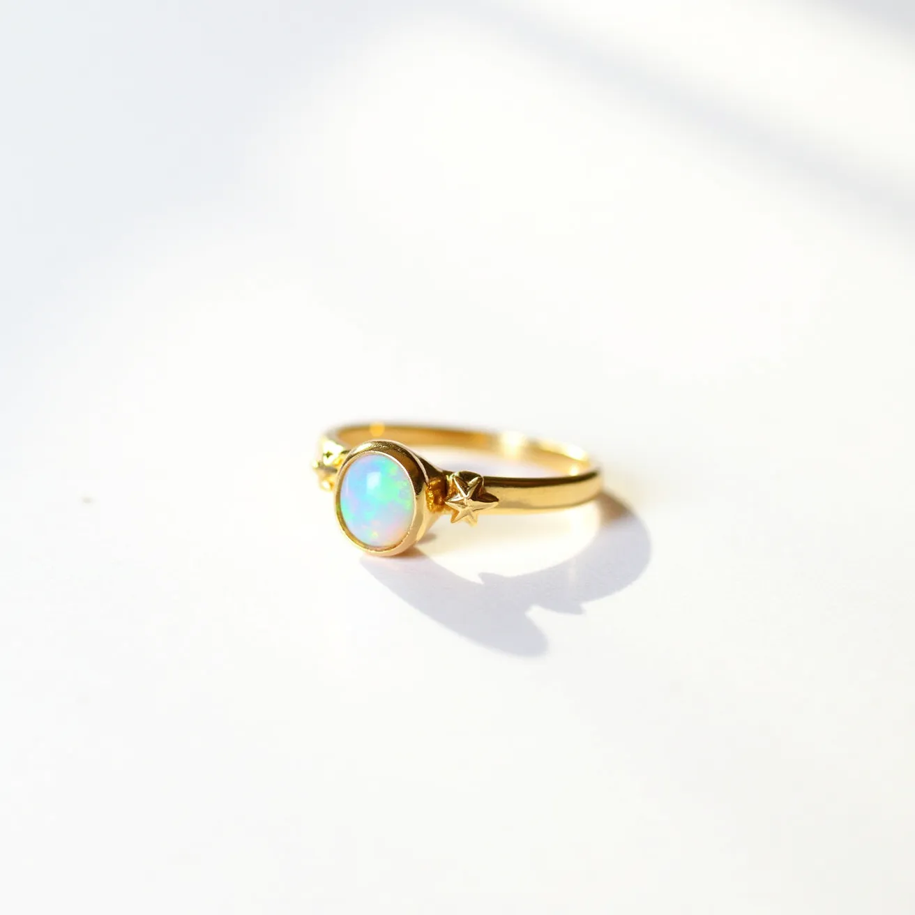 This opal ring features a luminous opal gemstone with a smooth oval cabochon cut, set in a bezel setting that securely holds the stone while showcasing its vibrant play of colors. The ring is crafted from a glossy gold metal, adding a warm and luxurious contrast to the opal. On either side of the opal, there are small gold star embellishments that add a whimsical and celestial element to the design. The band is delicate yet sturdy, complementing the central opal and stars. The overall style is elegant and modern, suitable for both everyday wear and special occasions.