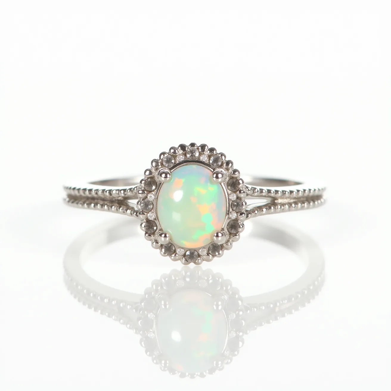 This opal ring features an elegantly crafted central opal gemstone that exhibits striking play-of-color with hues ranging from luminous greens to fiery oranges. The opal is oval-cut, providing a smooth, rounded presentation that enhances its natural iridescence. It is set within a delicate halo setting adorned with a series of smaller, rounded accent stones, possibly diamonds or cubic zirconia, adding a complementary sparkle. The band itself is made from a silver-toned metal, possibly white gold or sterling silver, with beaded detailing along its sides, contributing texture and sophistication to the design.