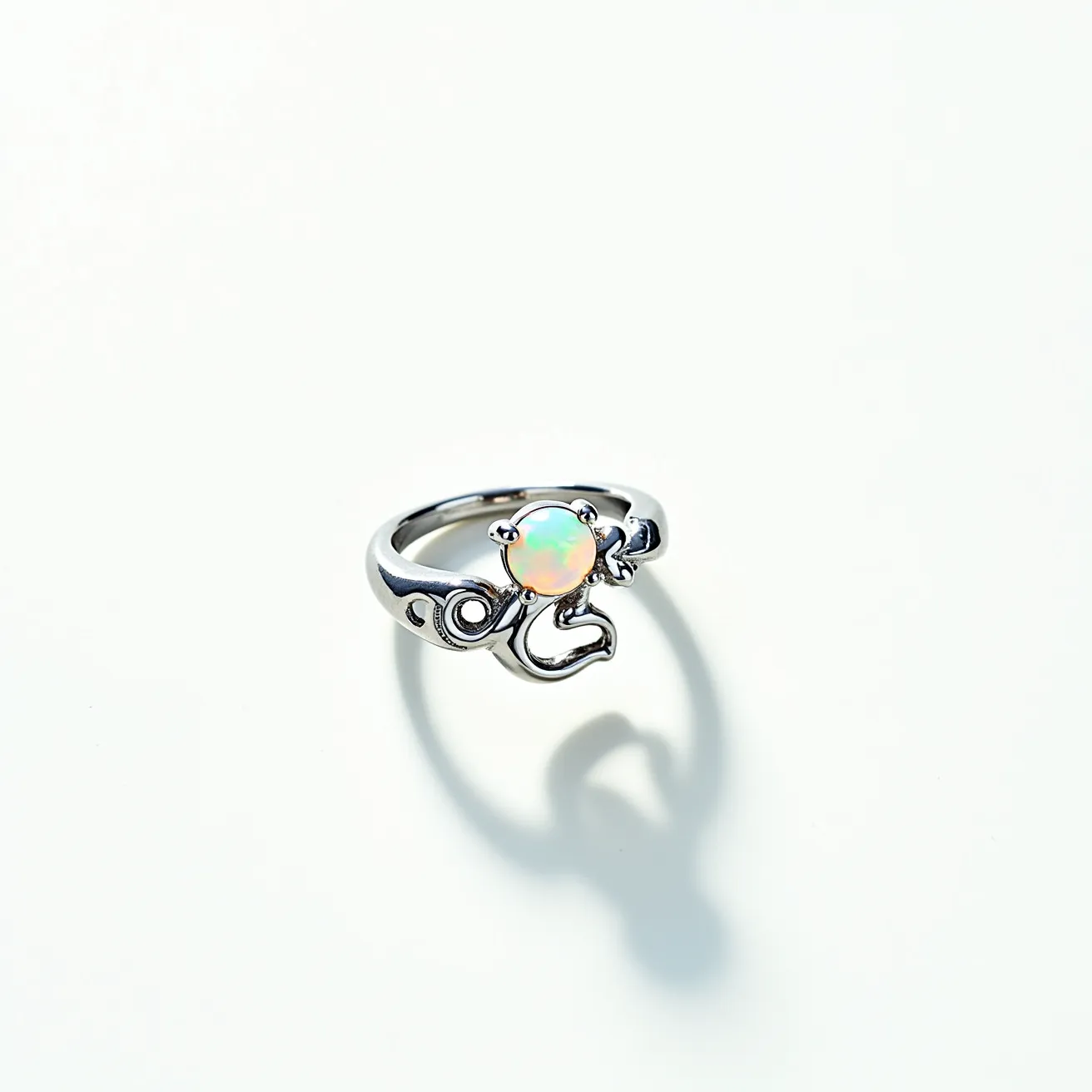 This opal ring features a central round opal gem with an iridescent play of colors, ranging from subtle greens to warmer hues, showcasing its classic beauty. The opal is securely set in a prong setting, allowing maximum exposure to light, enhancing its vibrant display. The band appears to be crafted from a polished metal, likely silver or white gold, which provides a sleek, reflective finish that complements the opal's glow. The ring design includes intricate, swirling elements on either side of the opal, adding a touch of elegance and uniqueness to the piece. The band is smooth and rounded, providing both comfort and a classic aesthetic.