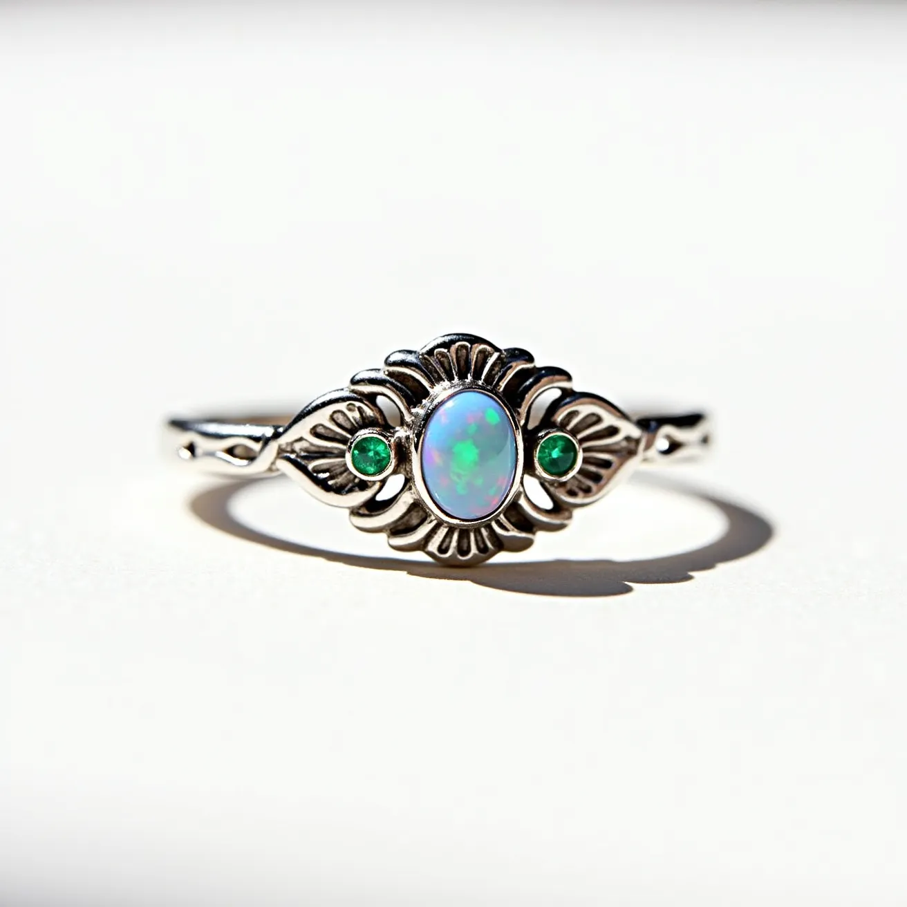 This opal ring features a central oval-shaped opal, which displays a captivating play of colors characteristic of this gemstone. Flanking the opal, two small, round, green gems complement its hues. The metal setting and band showcase an intricate design with a decorative pattern that enhances the elegance of the piece. The opal is securely held in place by a bezel setting, ensuring both protection and aesthetic appeal. This artistic design suggests craftsmanship that highlights both the beauty of the gemstones and the intricacy of the metalwork.