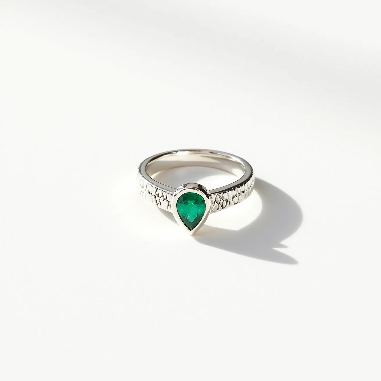 This open ring features a gleaming silver band with intricate, textured patterns along its surface, adding an elegant touch to its design. At the forefront of the ring is a pear-shaped green gemstone, likely an emerald, which commands attention with its vivid color. The gemstone is set in a bezel setting, securely encasing it and highlighting its contours. The overall presentation and materials suggest a blend of classic elegance and modern craftsmanship, making this piece both sophisticated and visually captivating.