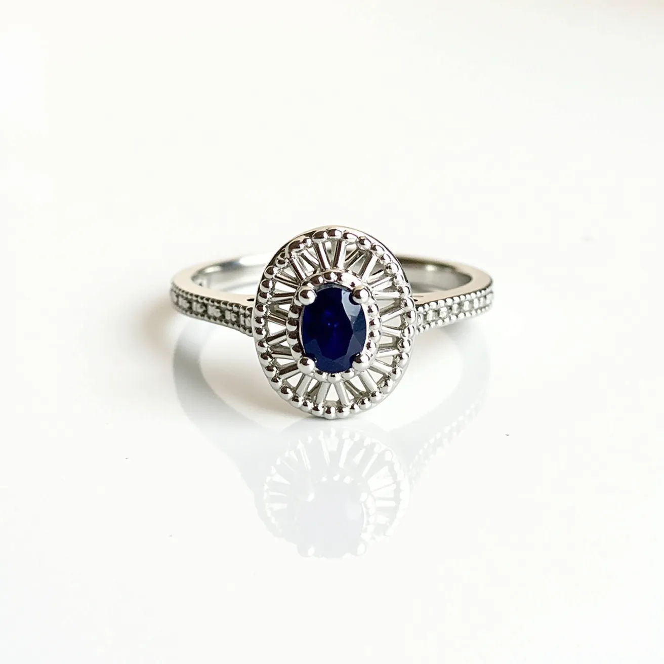 This oval engagement ring showcases an elegant design featuring a central dark blue gemstone, likely a sapphire, cut in an oval shape. The gemstone is encased in a detailed metal setting with a lattice design that radiates outwards, enhancing its sophisticated appearance. The band of the ring is adorned with small, sparkling accent diamonds or cubic zirconias, adding a refined touch of brilliance. The metal used for the band and setting appears to be a polished, reflective white metal, such as white gold or platinum, contributing to its luxurious feel. The design skillfully combines the vibrant centerpiece with delicate detailing, making it a striking piece.