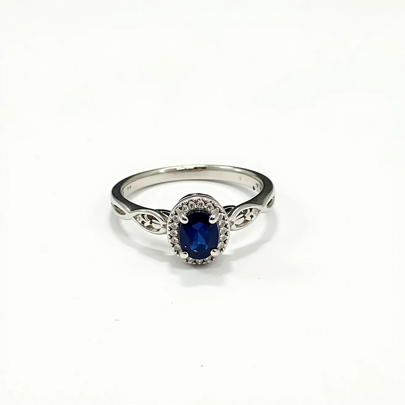 This oval engagement ring features a vibrant, deep blue oval gemstone, likely a sapphire, set at its center. The gemstone is surrounded by a halo of small, sparkling diamonds, enhancing its brilliance and creating a classic look. The main stone is securely held with a four-prong setting, which elevates the gemstone for maximum visibility. The band appears to be crafted from a silver-hued metal, possibly white gold or platinum, with delicate, intertwined designs on either side of the center stone, adding an element of sophistication and elegance. These decorative patterns transition smoothly into the simple, polished band that completes the ring's graceful aesthetic.