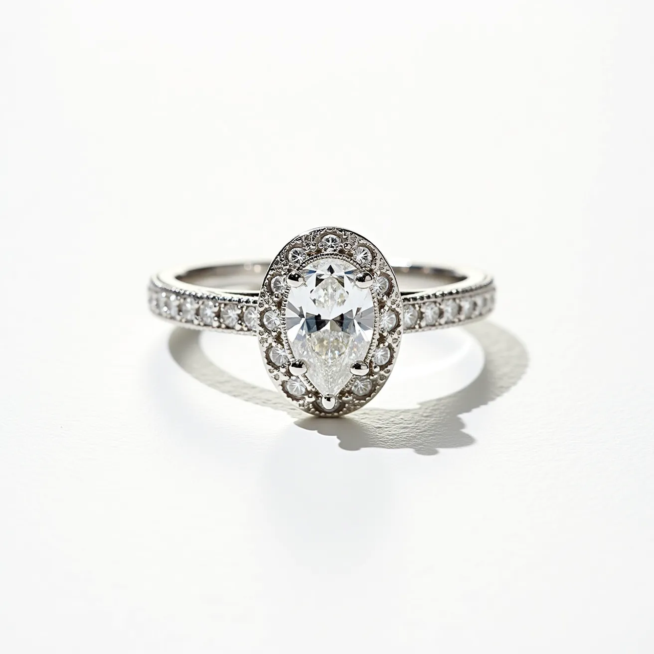 This oval engagement ring features a central pear-shaped diamond, elegantly set in a halo of smaller round diamonds that enhance its brilliance. The band is crafted from a polished metal, most likely white gold or platinum, known for its durability and lustrous appearance. Additional round diamonds are set into the band, adding a continuous sparkle that complements the intricate design of the centerpiece. The prongs securely hold the pear-shaped diamond, highlighting its clarity and cut, while the halo setting adds an extra layer of opulence to this sophisticated piece.