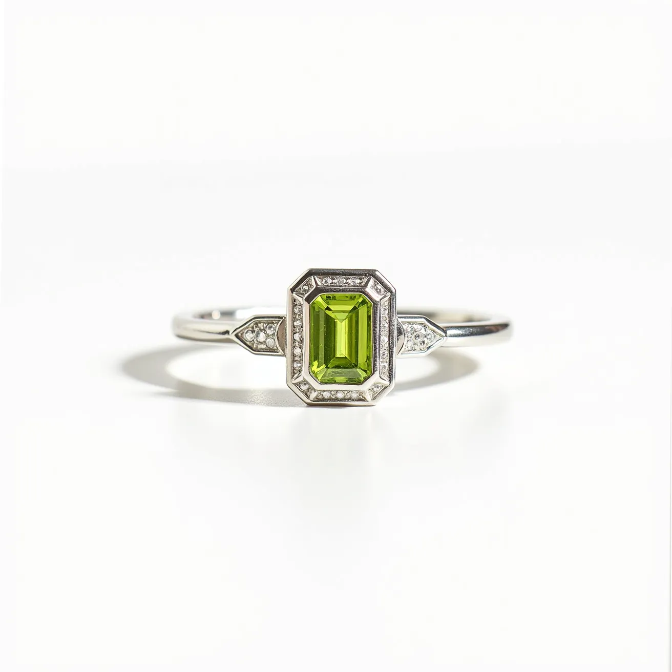 This peridot engagement ring features an emerald-cut peridot gemstone as its centerpiece, exhibiting a vibrant green hue. The peridot is set in a bezel setting, surrounded by a halo of small, sparkling diamonds that enhance its brilliance. The band appears to be made of polished white gold or platinum, contributing to the ring's elegant and refined look. On either side of the peridot, the shoulders of the ring are adorned with additional small diamonds arranged in a geometric design. The combination of the vivid peridot and the shimmering diamonds creates a striking and timeless piece.