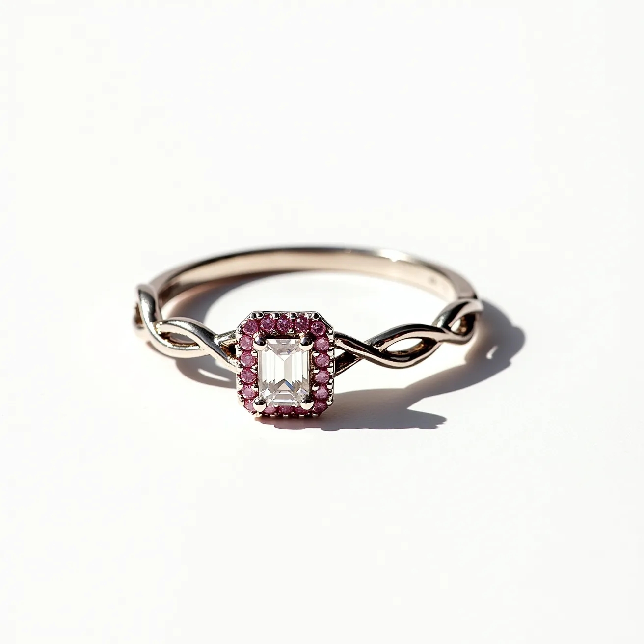 This pink diamond ring features a central emerald-cut diamond set within a halo of small pink gemstones, possibly pink sapphires. The band is crafted from a polished metal, likely white gold or platinum, with an elegant twisted design that adds a touch of sophistication to the overall aesthetic. The halo setting enhances the brilliance of the central diamond, while the pink stones provide a charming contrast. The seamless integration of the materials and stones creates a harmonious balance, making it a striking piece of jewelry.