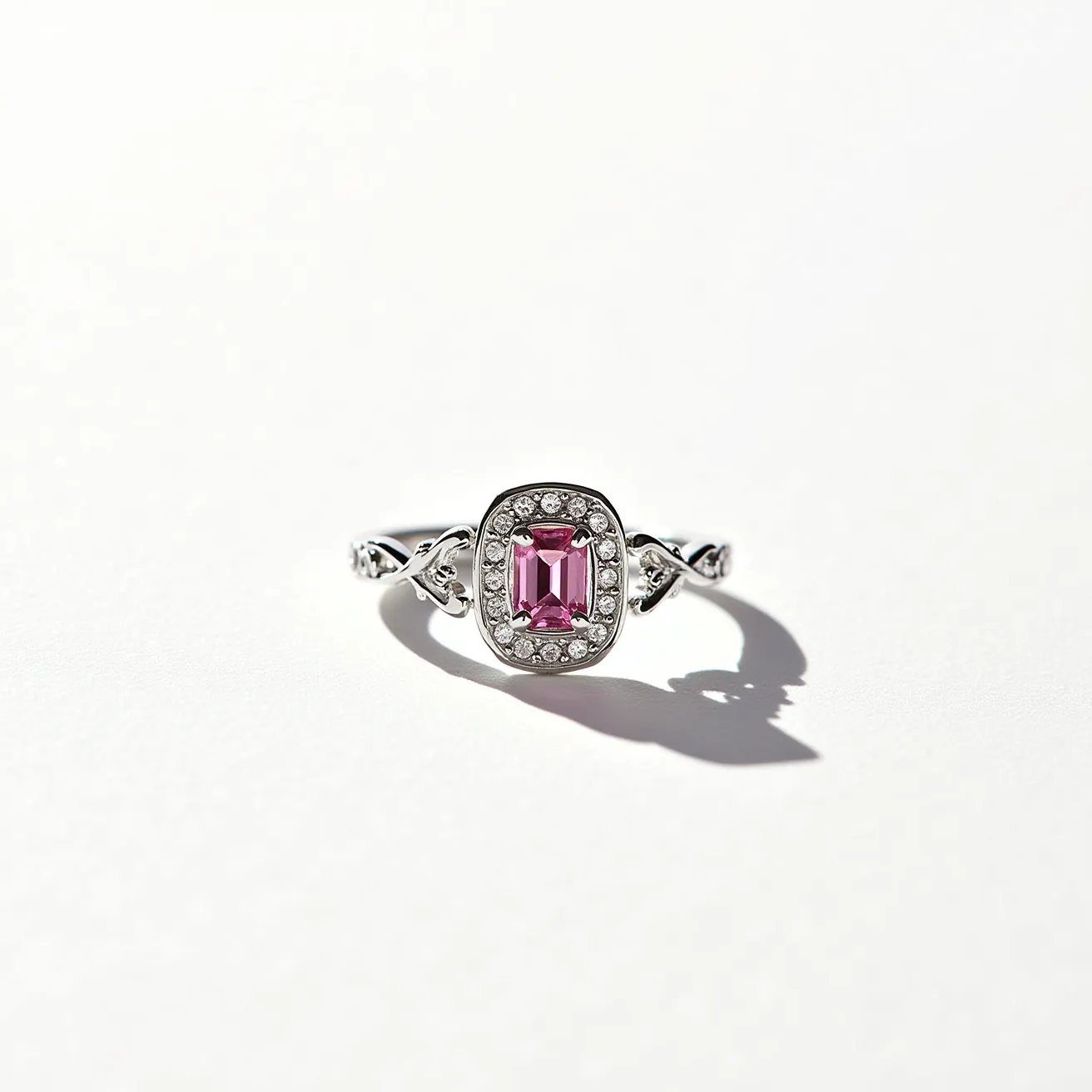 This pink diamond ring features a captivating, emerald-cut pink diamond as its central stone, elegantly set in a prong setting. The central gem is surrounded by a halo of smaller, round white diamonds, enhancing its vibrant hue and adding a touch of brilliance. The delicate band is crafted from polished white gold or platinum, adorned with subtle infinity motifs on either side, adding an element of sophistication and design complexity. The overall craftsmanship of the ring highlights the exquisite contrast between the pink and white gems, making it a stunning piece of jewelry.